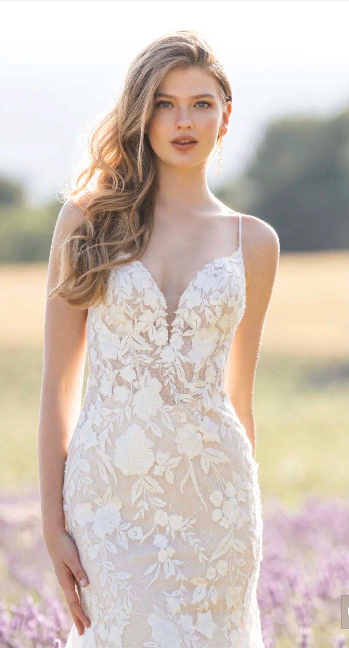 A person wearing a white, floor-length, fit-and-flare wedding dress with a deep V-neckline, standing outdoors.