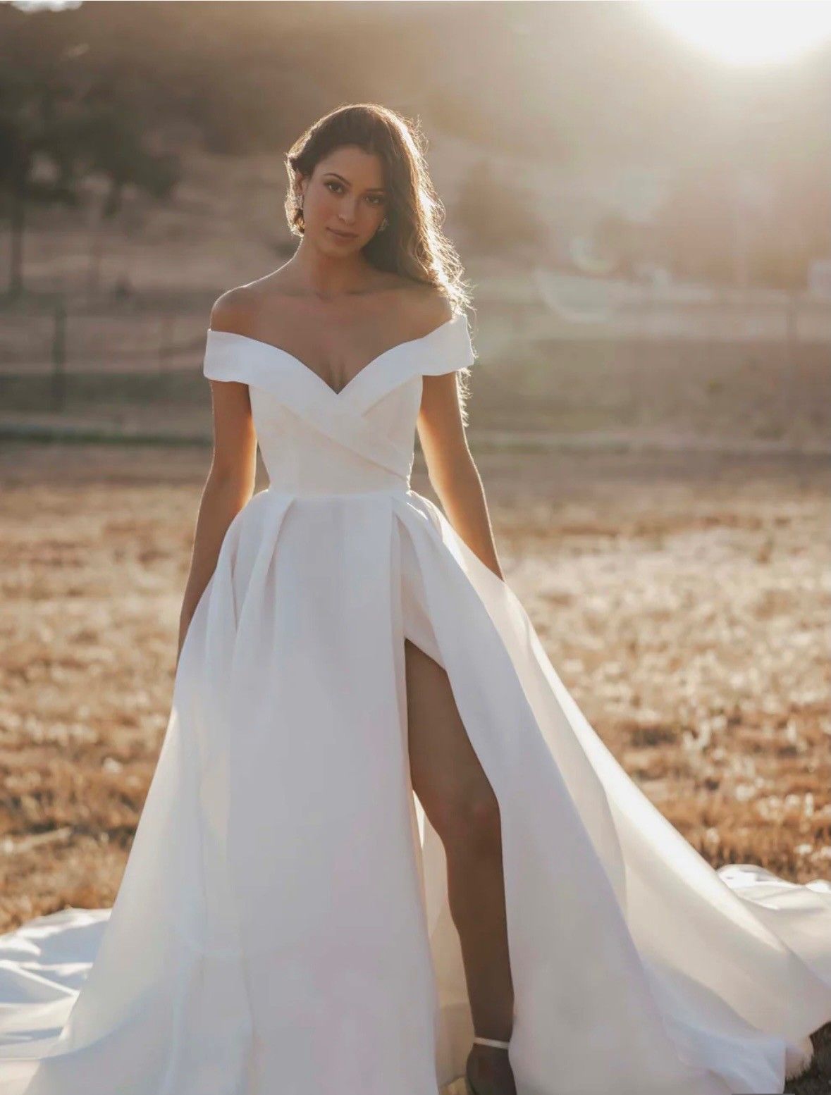 A person wearing a white, off-the-shoulder wedding dress with a high leg slit, standing in a field at sunset.