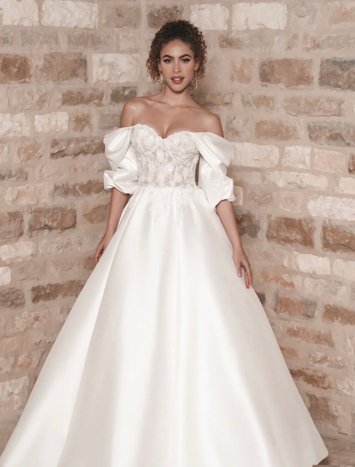 A person wearing an off-the-shoulder, white A-line wedding gown with a floral lace bodice, standing against a brick wall.