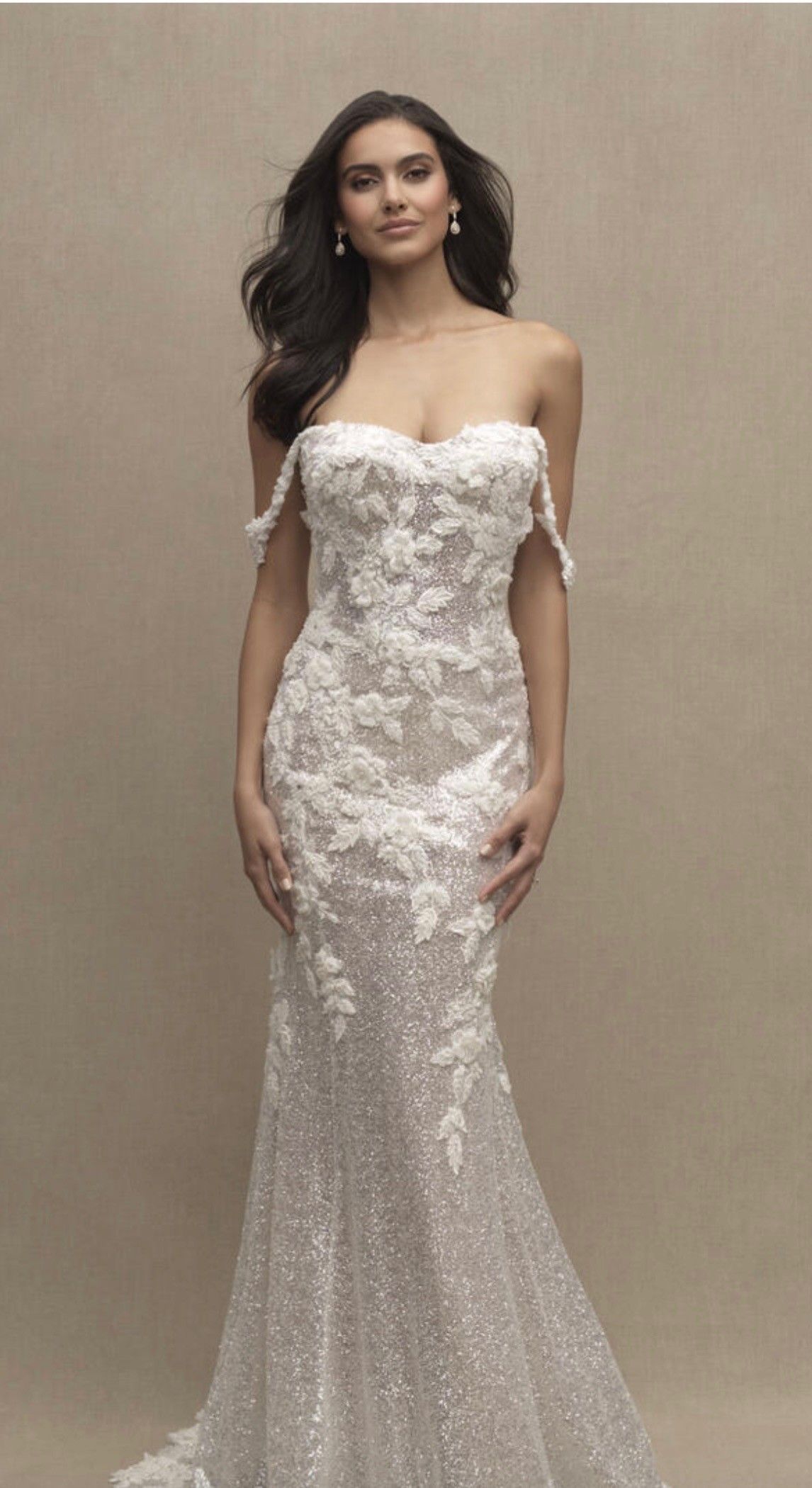 A person poses in a strapless, floor-length wedding gown featuring intricate floral lace appliqué and a sequined skirt.