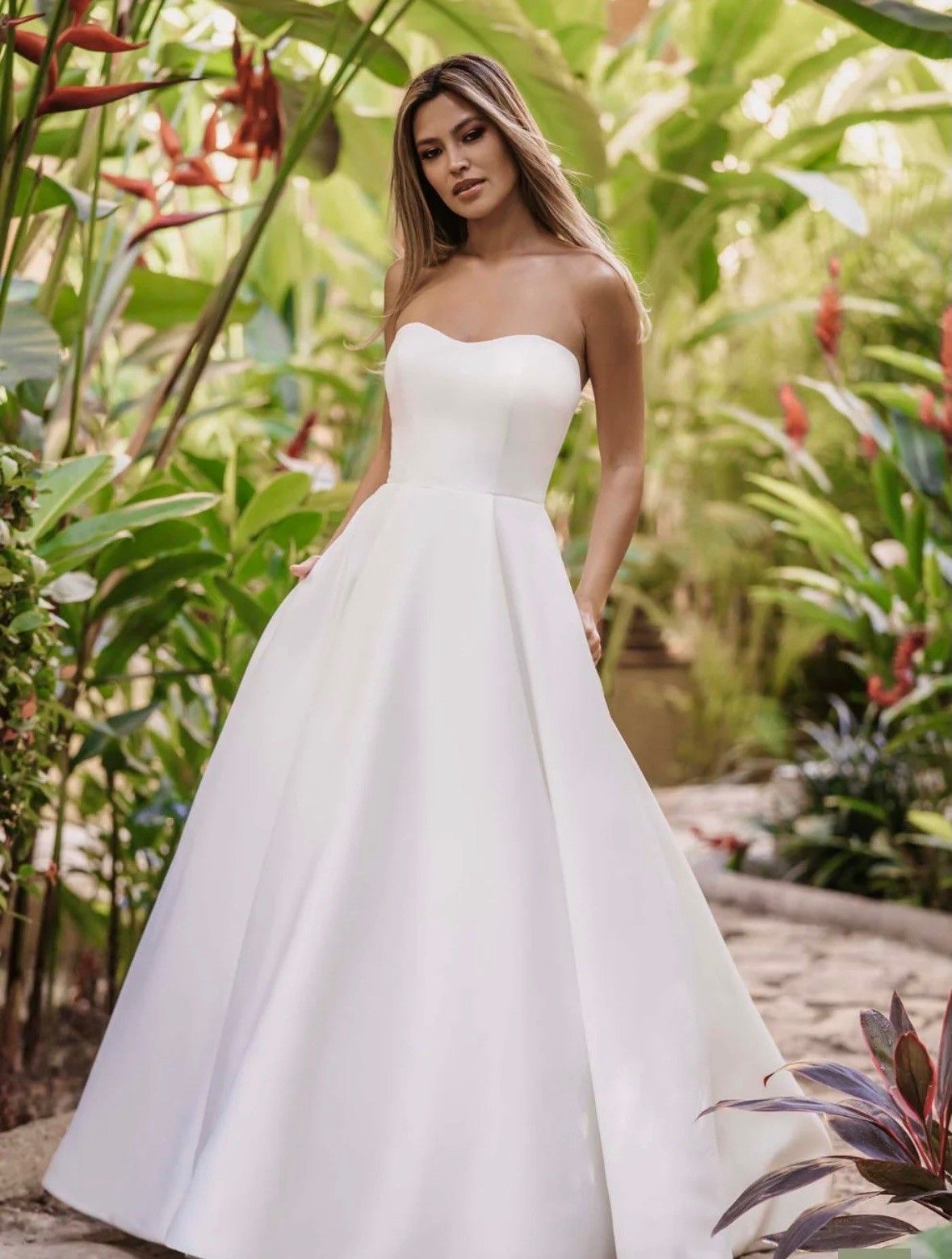 A person wearing a white, strapless, A-line wedding dress posing in a lush, green tropical garden.