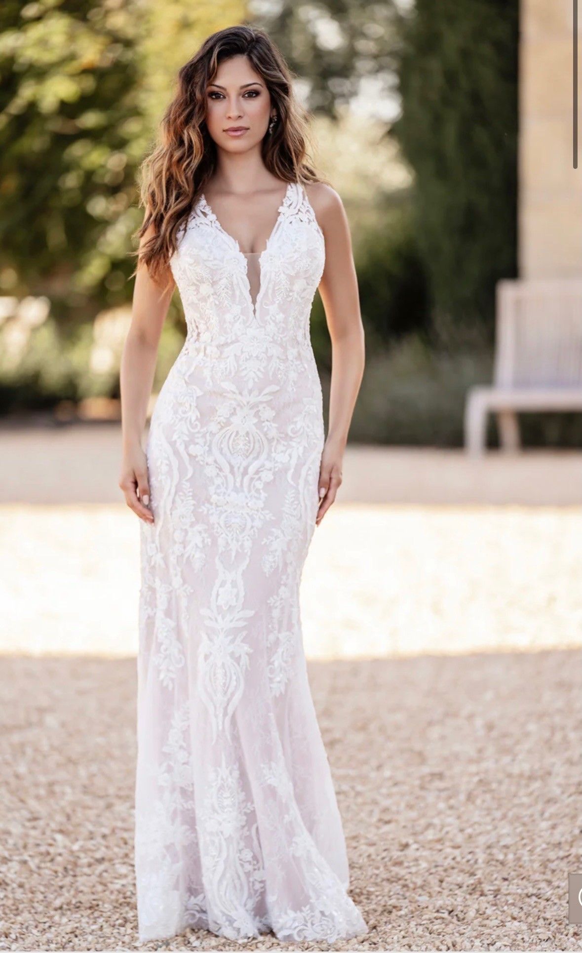 A person standing outdoors wearing a white, floor-length lace wedding dress with a deep V-neckline.