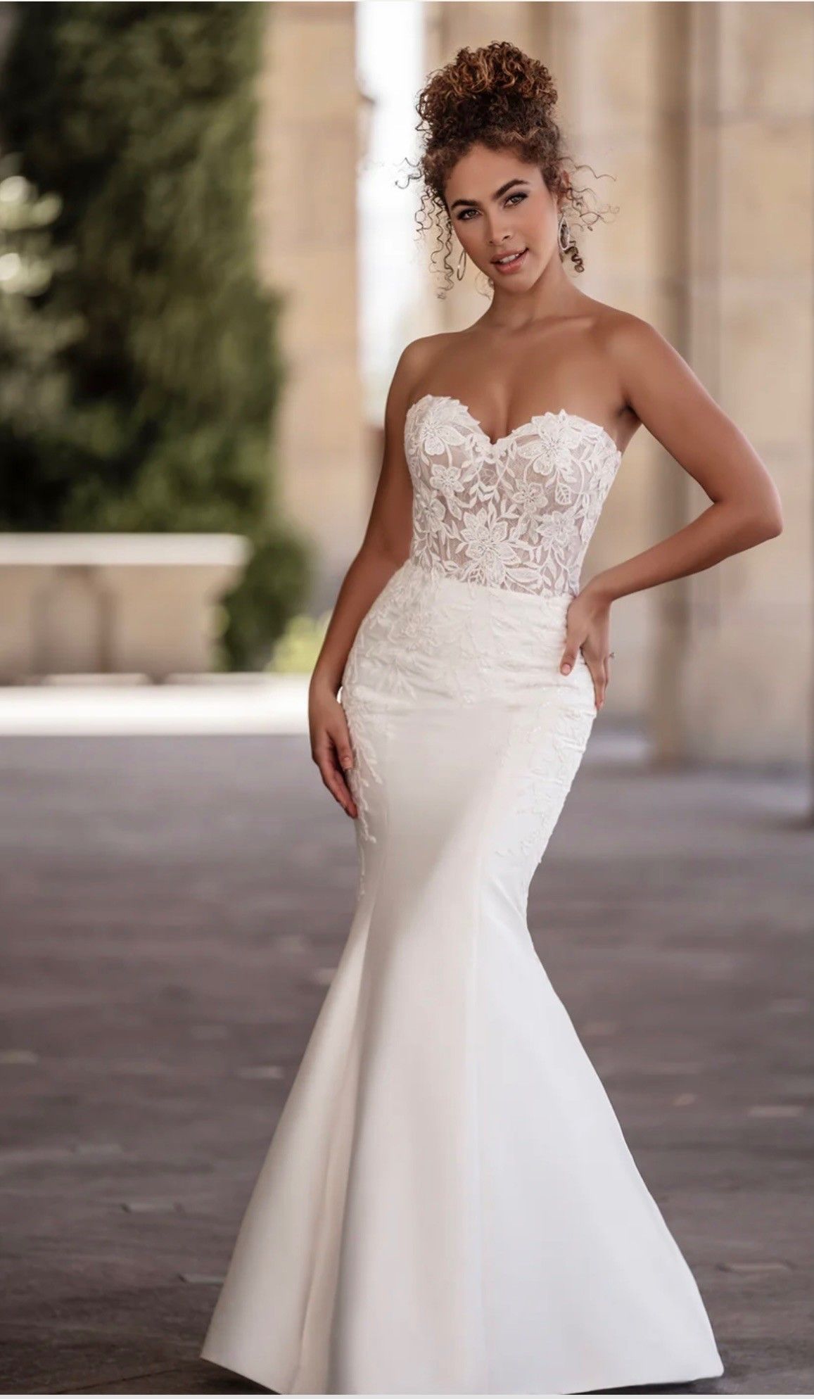 A strapless wedding dress with a lace bodice and a floor-length, fitted satin skirt, worn by a person posing outdoors.