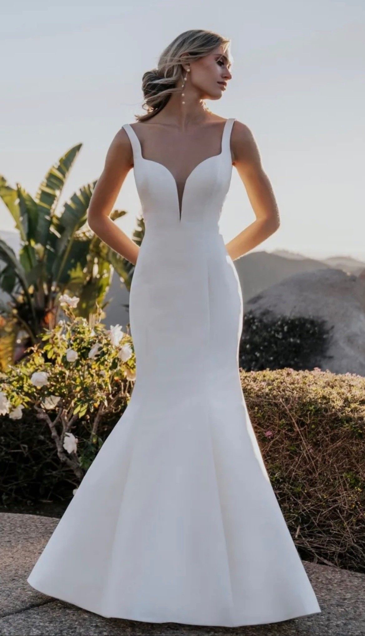 A person wearing a white, floor-length, fit-and-flare wedding dress with a deep V-neckline, standing outdoors.