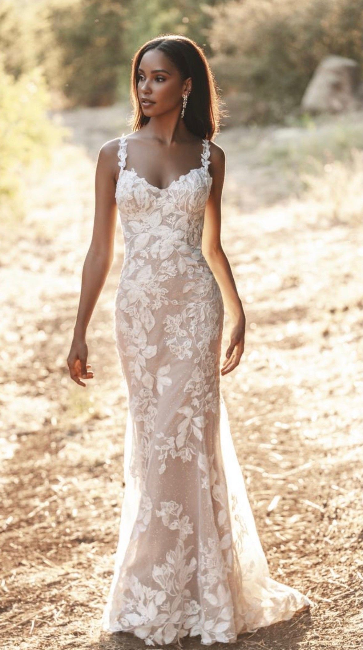 A person poses outdoors in a sleeveless, floor-length wedding gown with a white floral lace overlay on a nude lining.