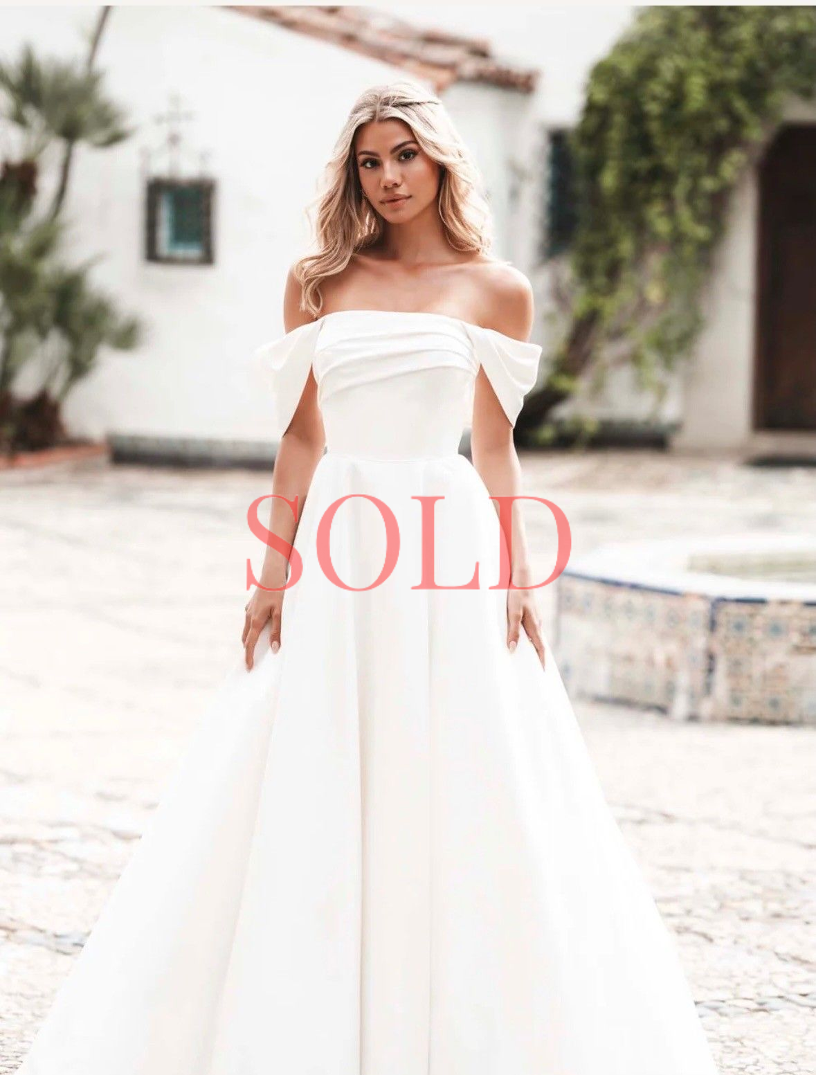 A person stands in a sunlit courtyard wearing an elegant, white off-the-shoulder wedding gown with a full skirt.