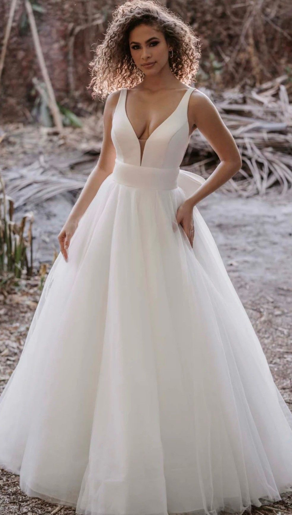 A person with curly hair wearing a white, V-neck A-line wedding gown with a tulle skirt, posing outdoors.