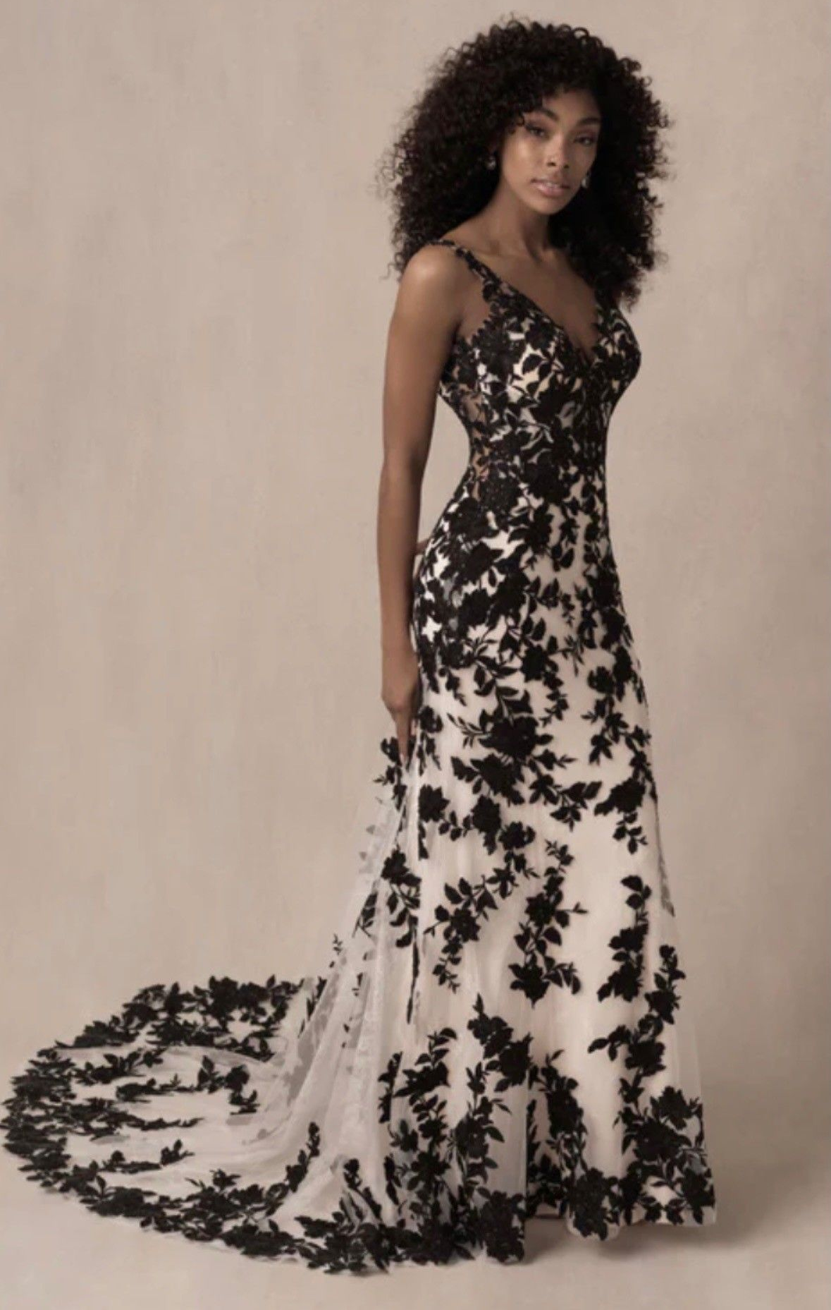 A full-length, A-line gown with black floral lace overlays on a cream background, featuring a V-neckline and a long train.