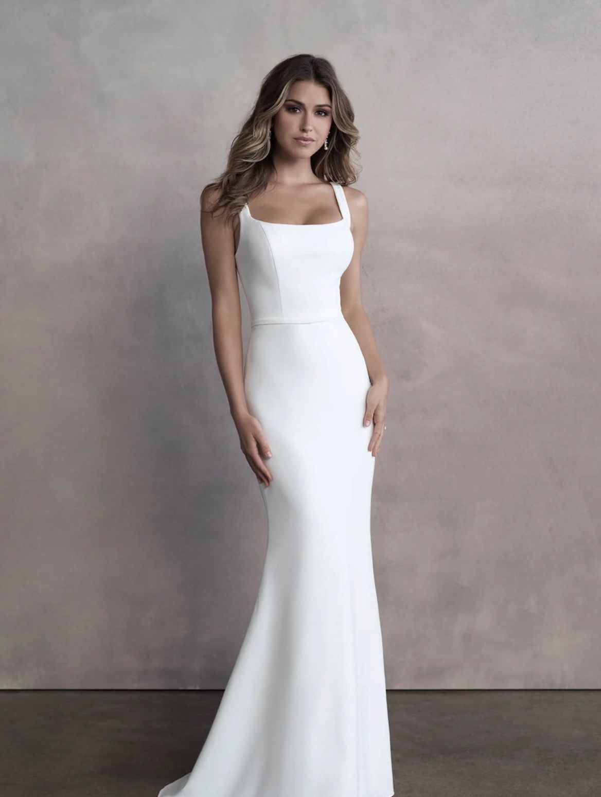 A person wears a minimalist white floor-length wedding gown with thick straps and a fitted silhouette against a grey wall.