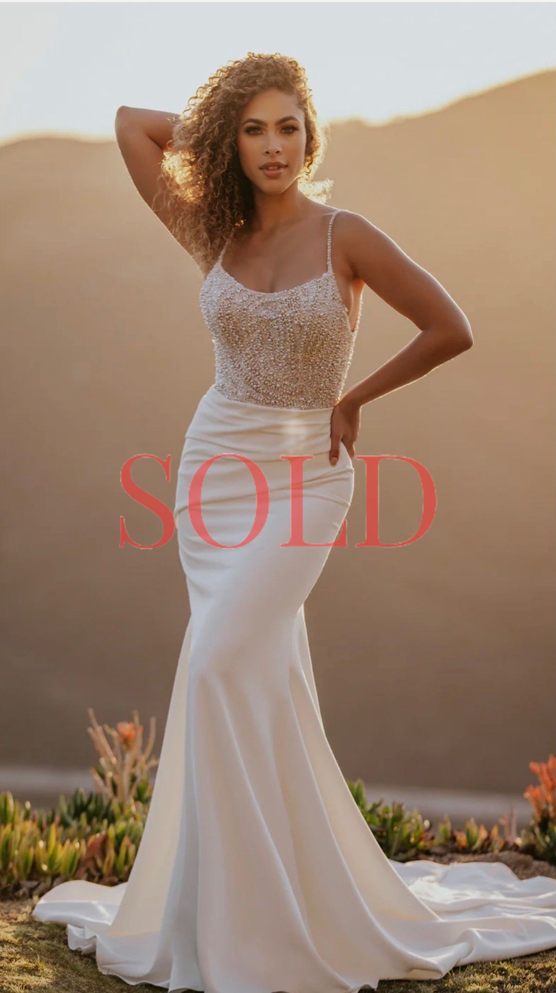 A person with curly hair wearing a white, beaded-bodice wedding dress, standing outside at sunset with a desert backdrop.