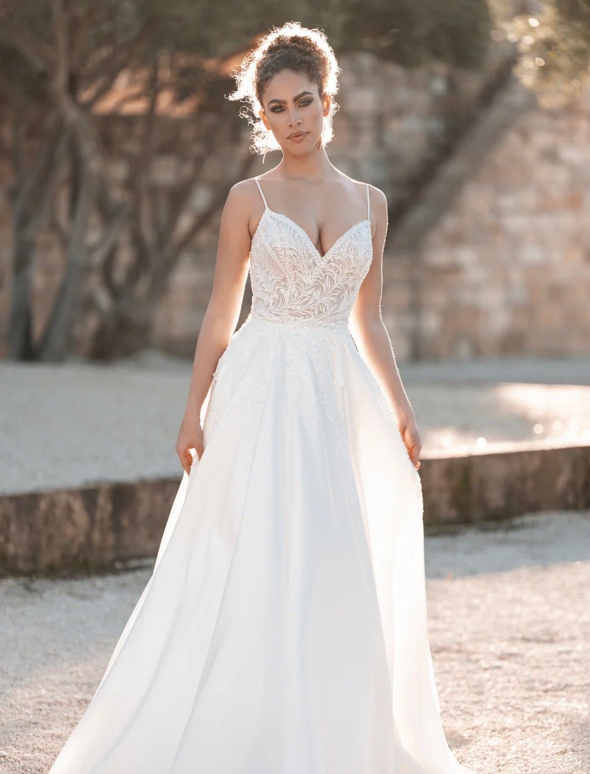 A model in a white lace-bodice wedding dress with thin straps stands outdoors against a blurry, sunlit background.