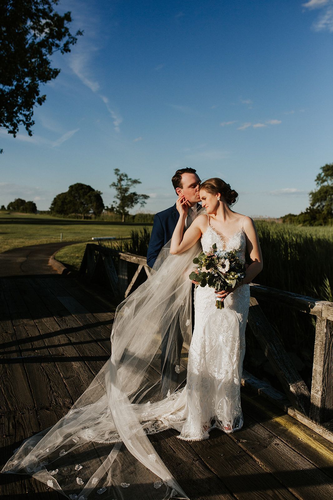 A bride and groom are kissing on a bridge . the bride is wearing a long veil.