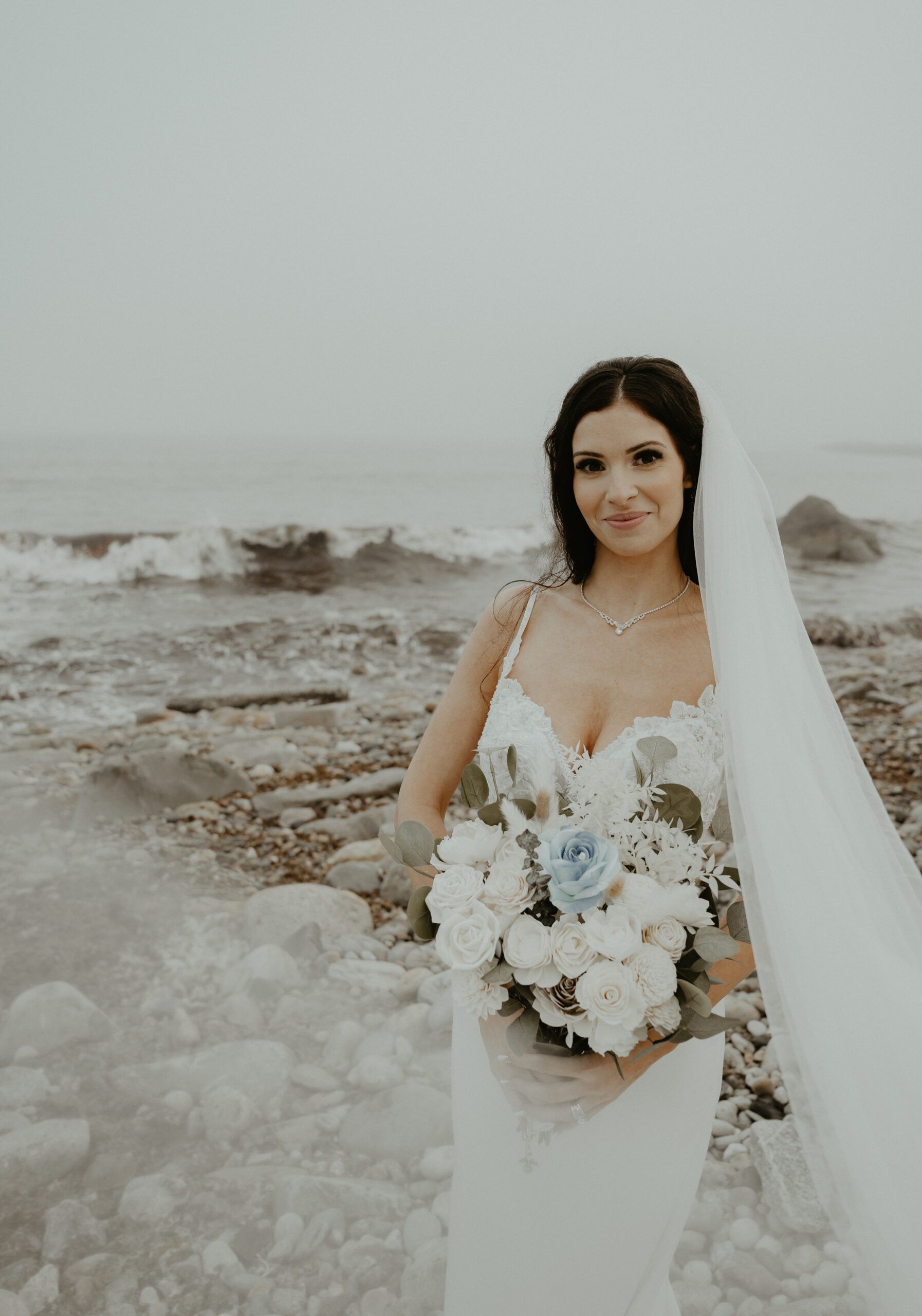 A bride in a wedding dress and veil is holding a bouquet of flowers on the beach.