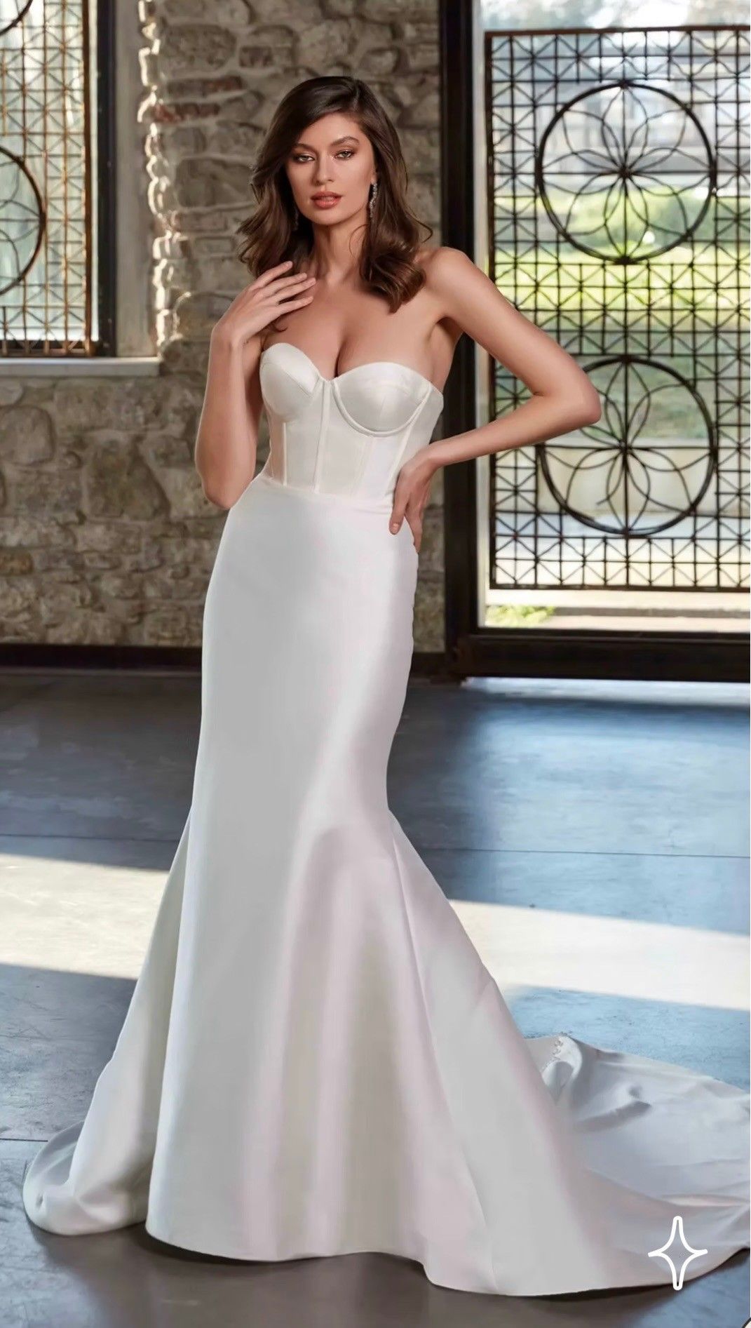 A person wearing a strapless, floor-length white satin mermaid wedding dress, standing in a room with stone walls.