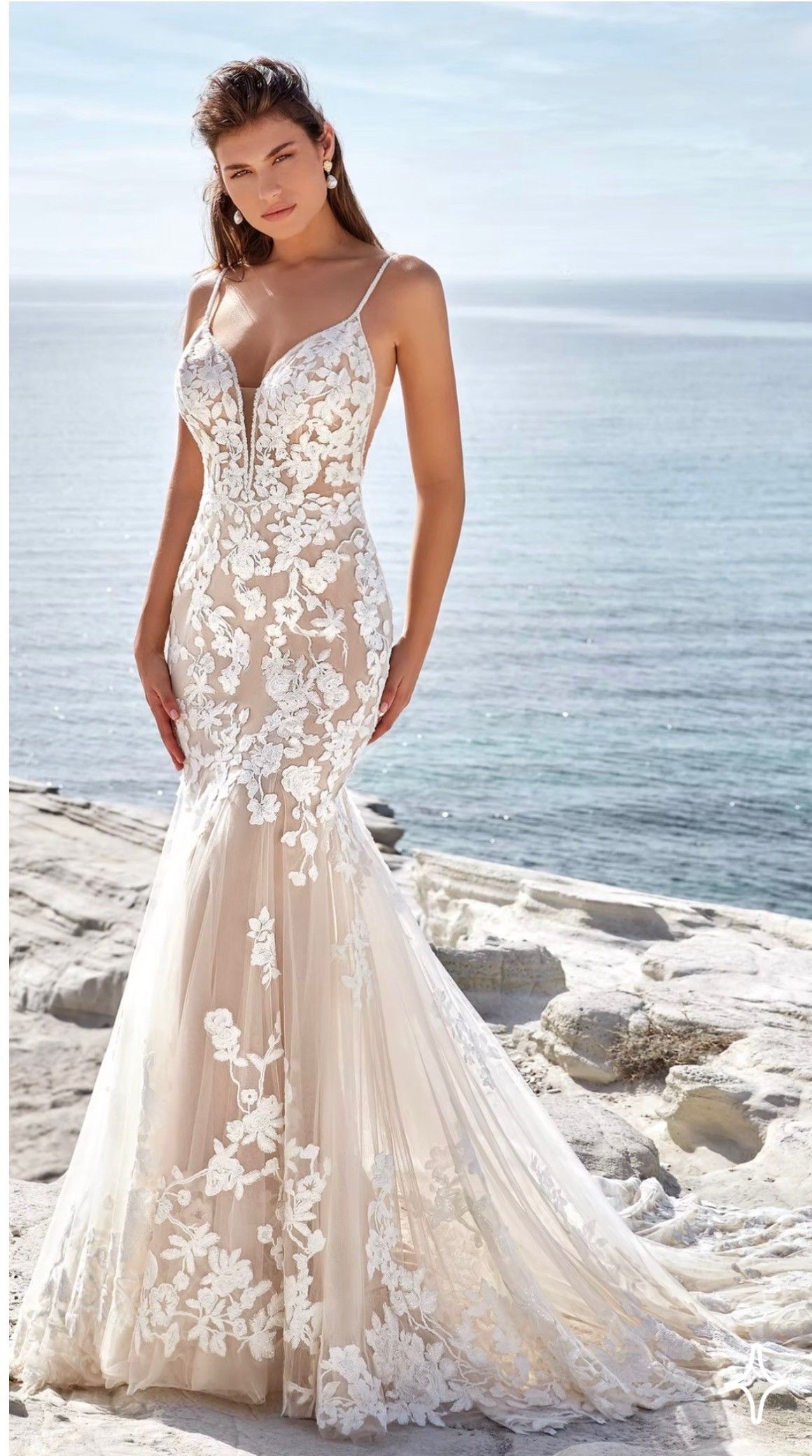 A model wears a fitted, white floral lace mermaid-style wedding dress on a rocky beach by the ocean.
