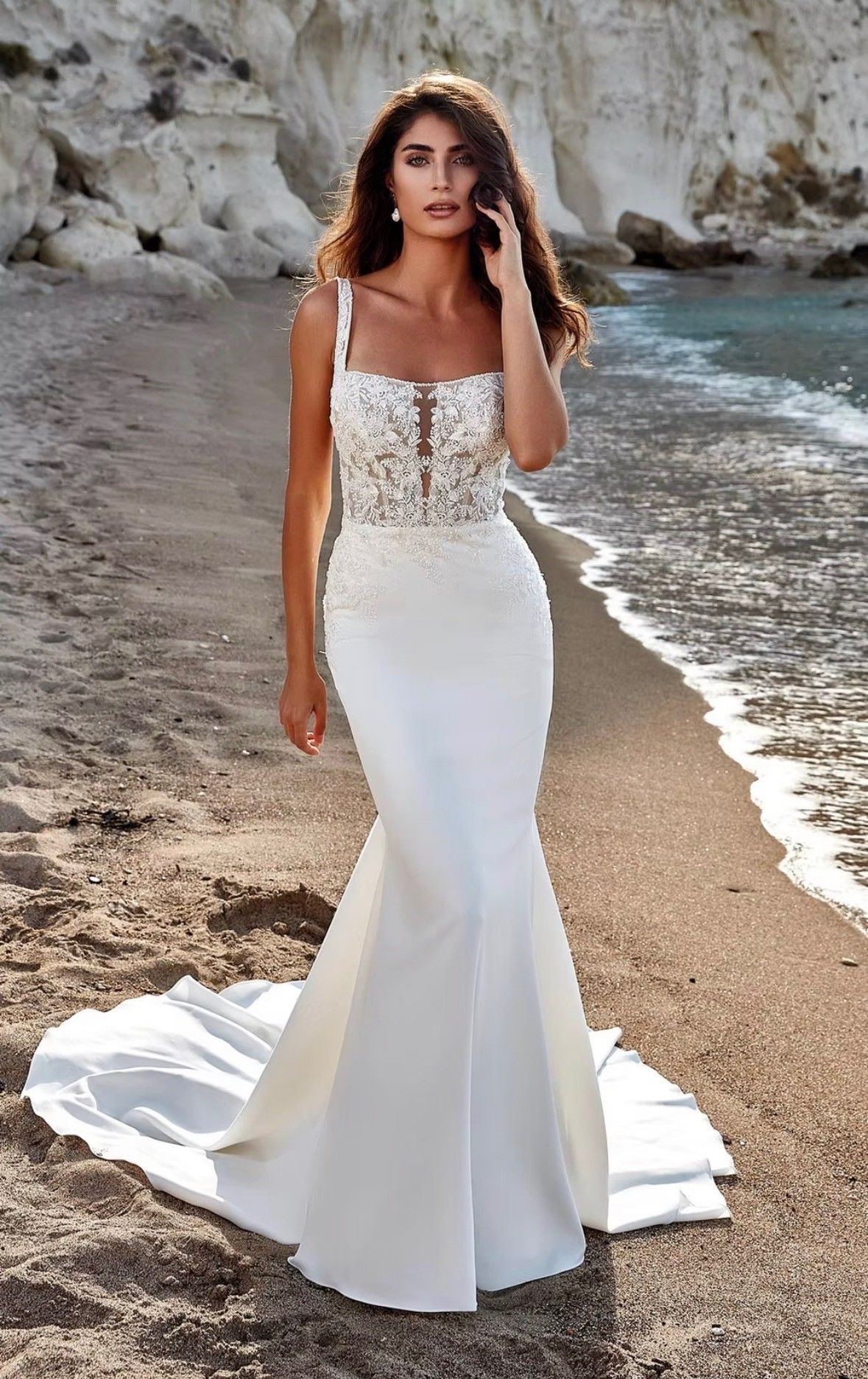 A woman models a white, floor-length mermaid bridal gown with a lace bodice on a sandy beach by the ocean.