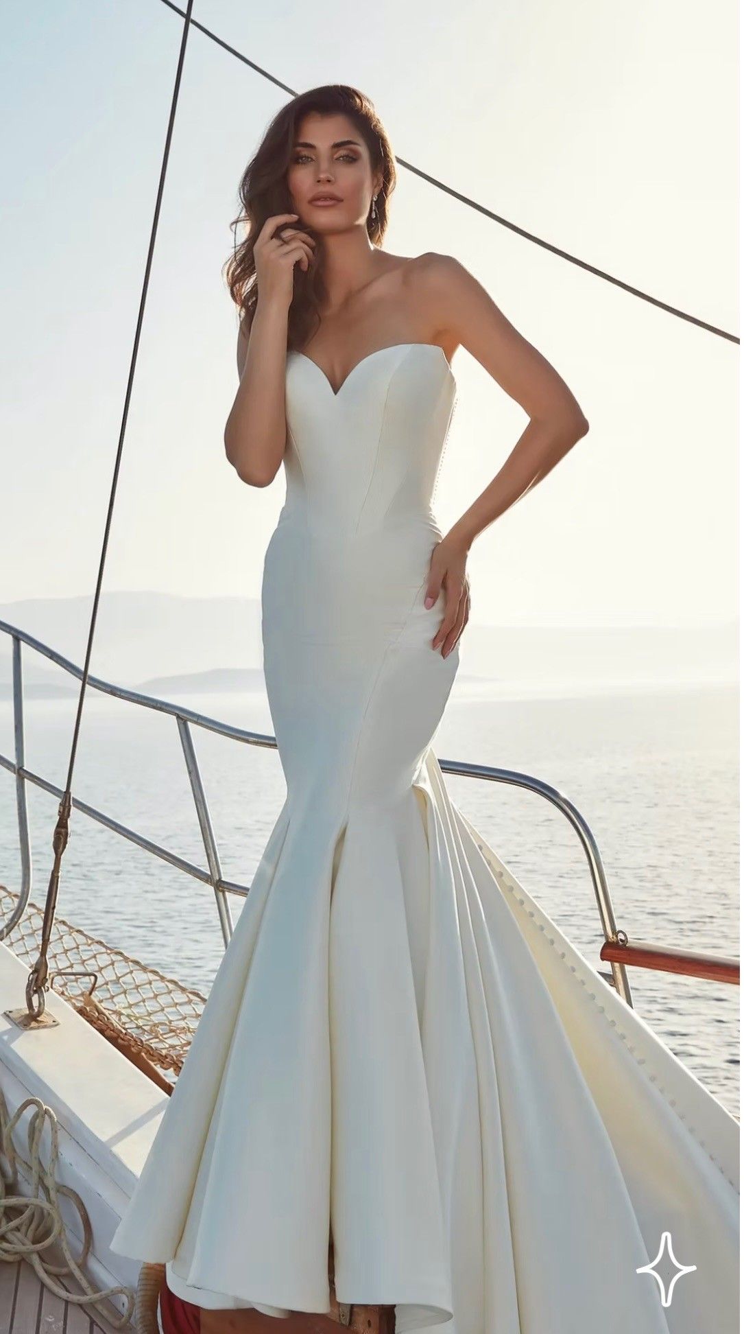A person stands on a boat wearing a strapless, floor-length white mermaid-style wedding gown with a front slit.