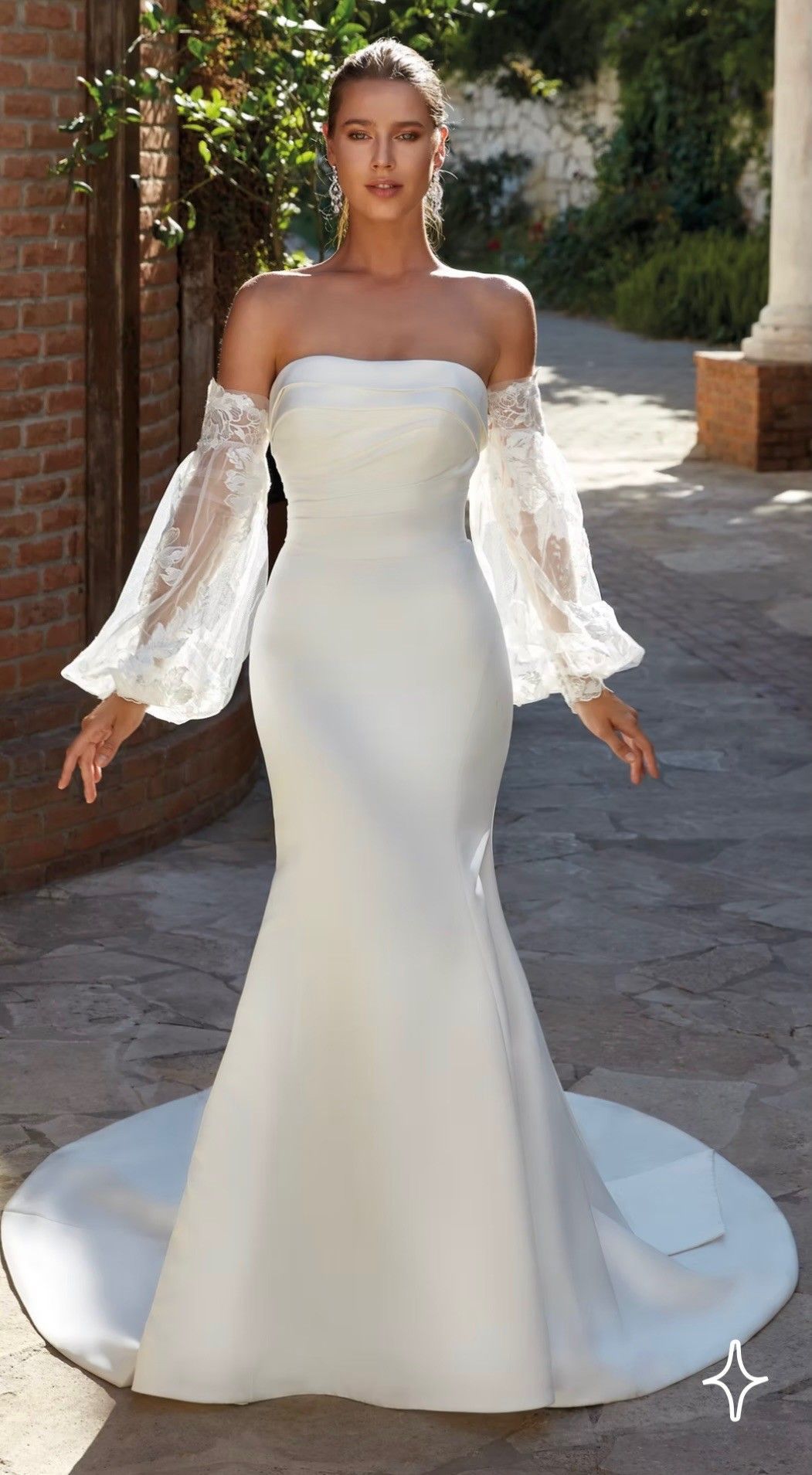 A person wearing a strapless, floor-length white wedding dress with detachable long lace bell sleeves, outdoors.