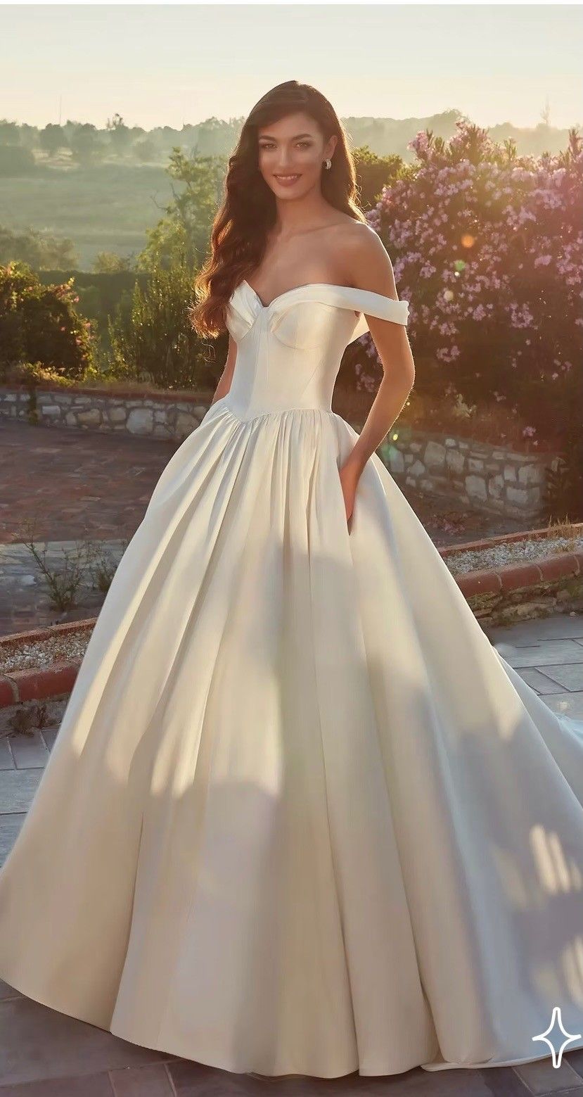 A model wearing an off-the-shoulder, white satin ballgown with a full, pleated skirt in a sunlit outdoor setting.