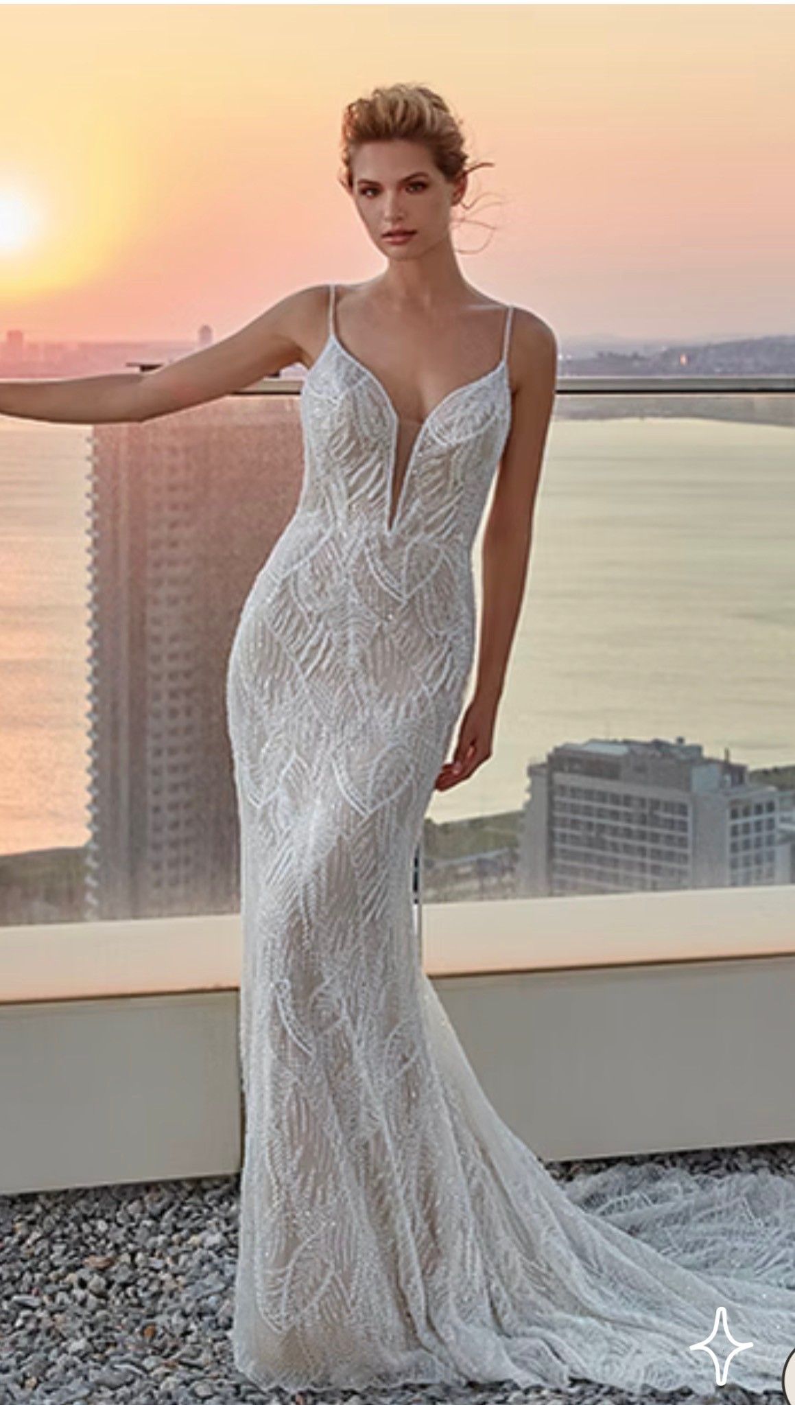 A person poses in a form-fitting, beaded white wedding dress with a deep V-neckline against a sunset city skyline.