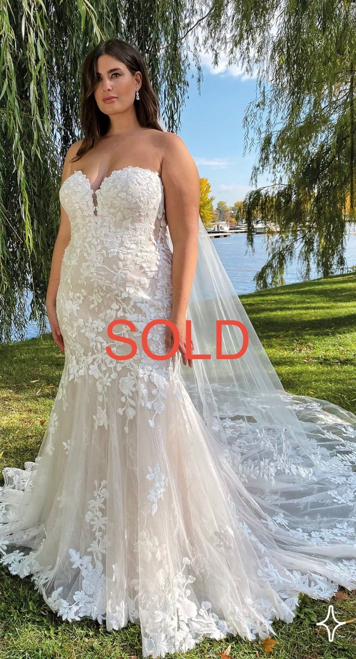 A person wearing a strapless, floral lace mermaid wedding dress with a long train, standing outdoors by a river.