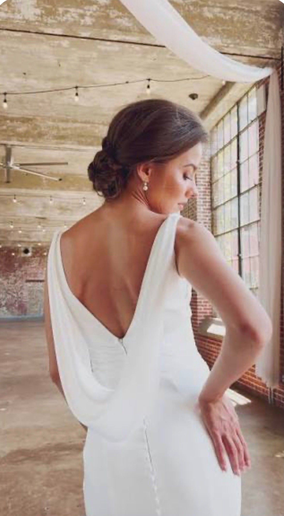 A person wearing a white, backless wedding gown with a draped cowl design, standing in a rustic, light-filled venue.