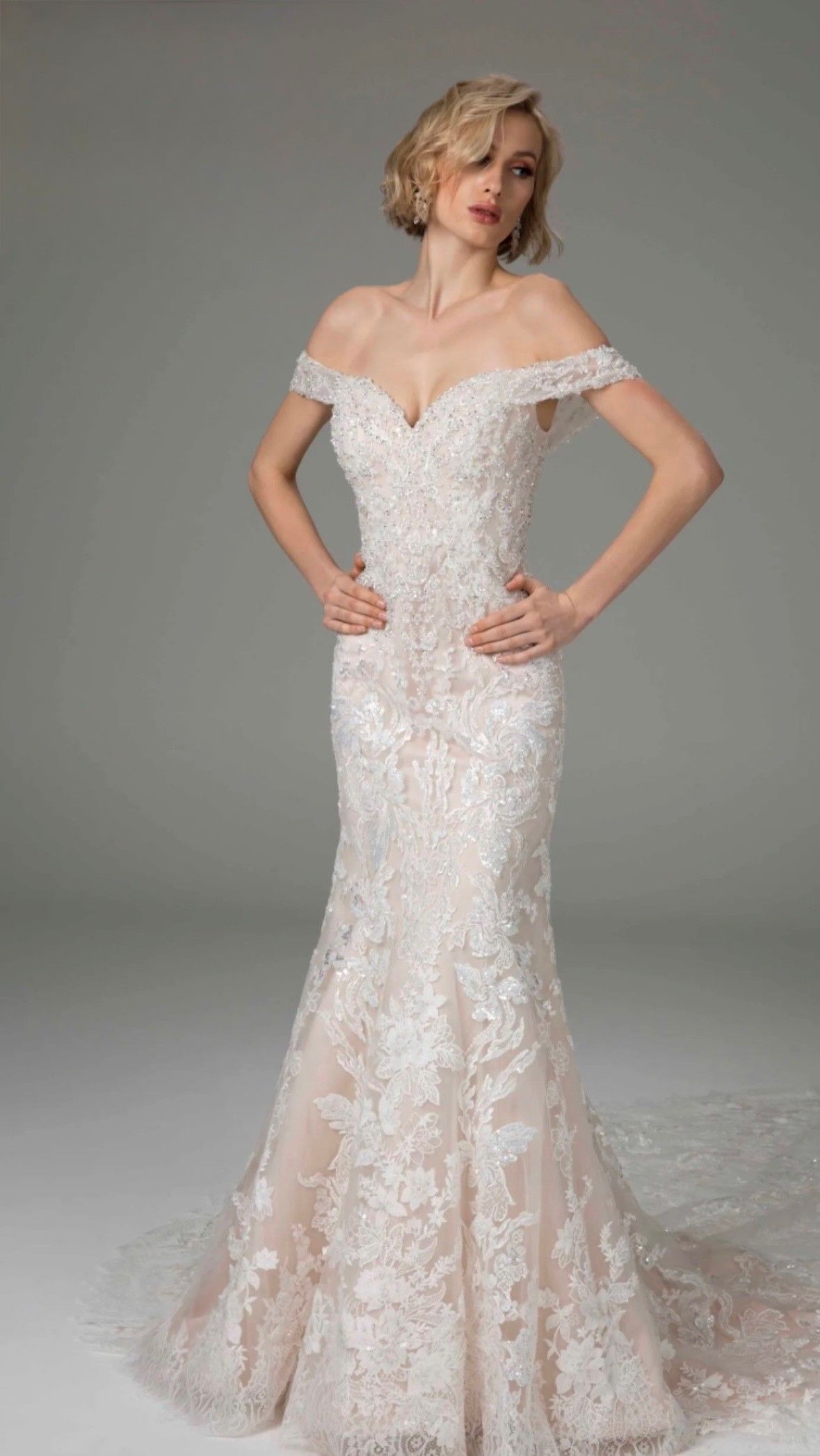 A blonde person wearing an off-the-shoulder, floor-length, white lace mermaid wedding dress against a plain gray background.