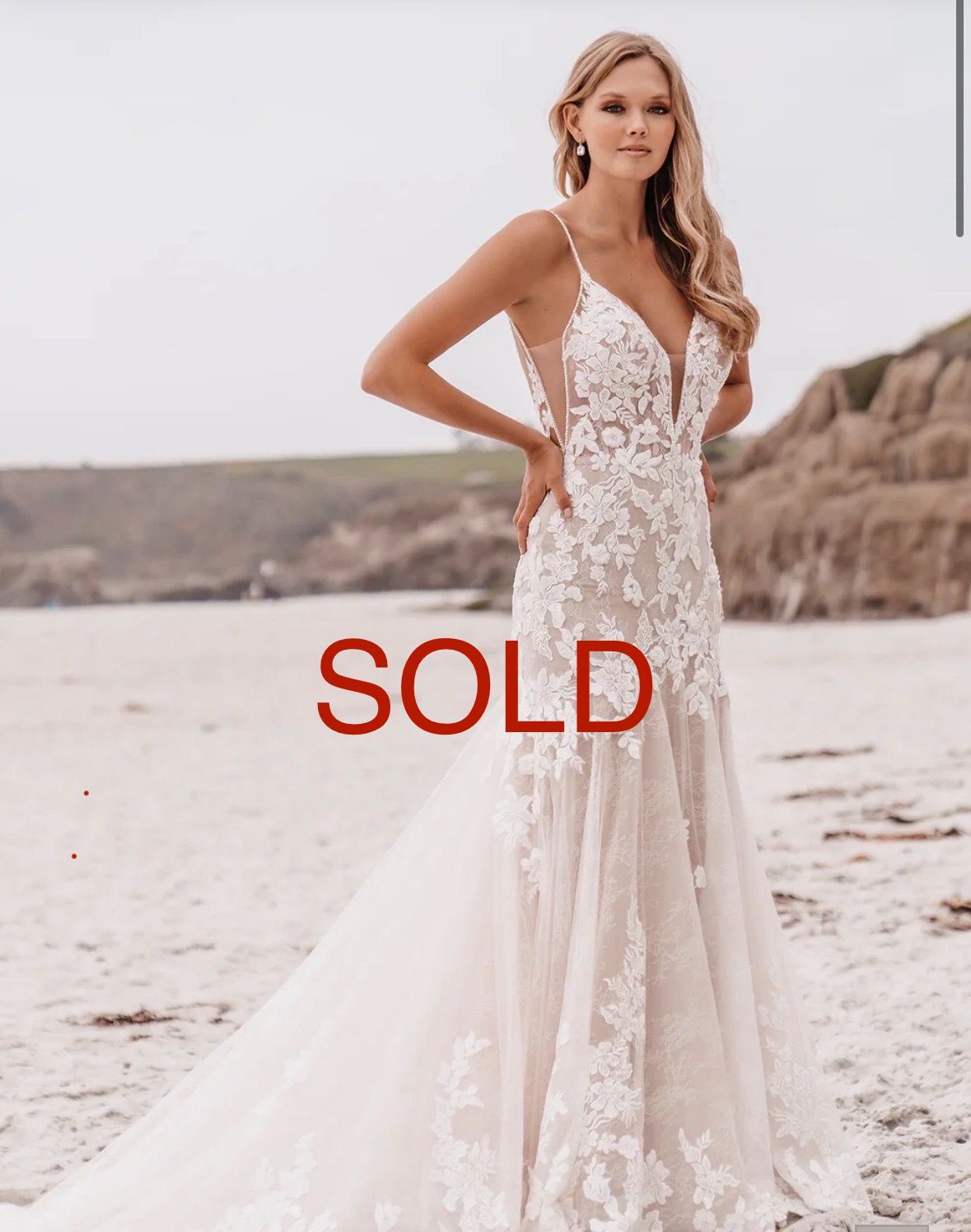 Woman in a wedding dress on a beach, smiling. She wears a white gown with lace and a long train.