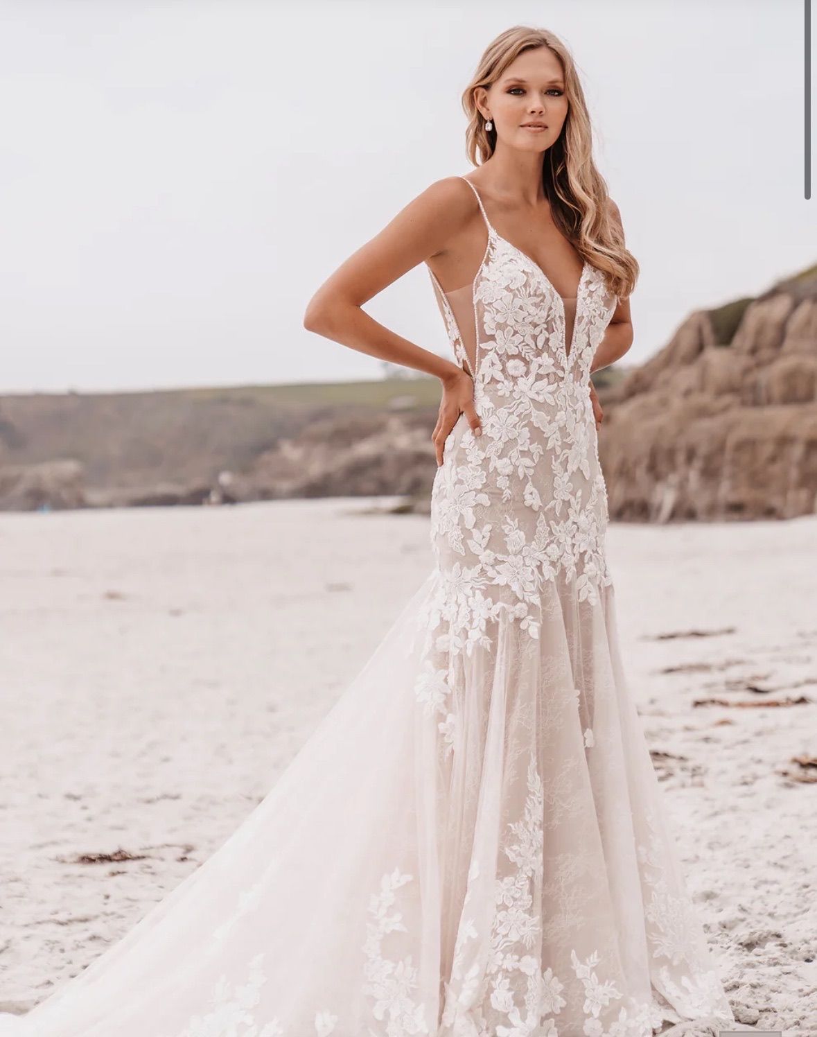 Woman in a wedding dress on a beach, smiling. She wears a white gown with lace and a long train.