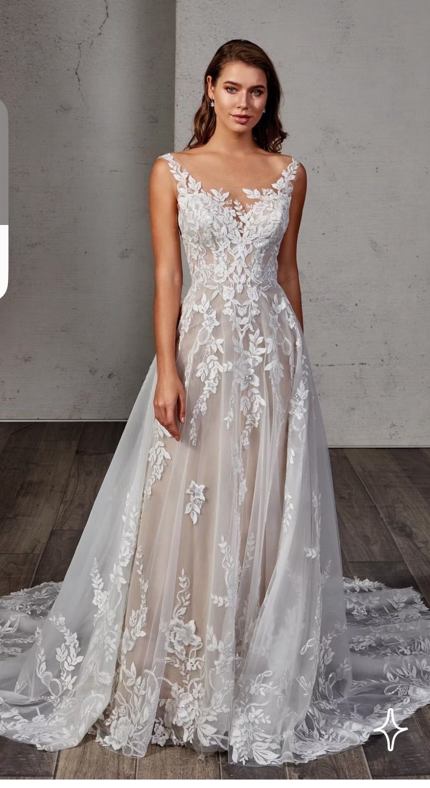 Woman in a white wedding dress with lace detail, standing on a wood floor with a textured wall.