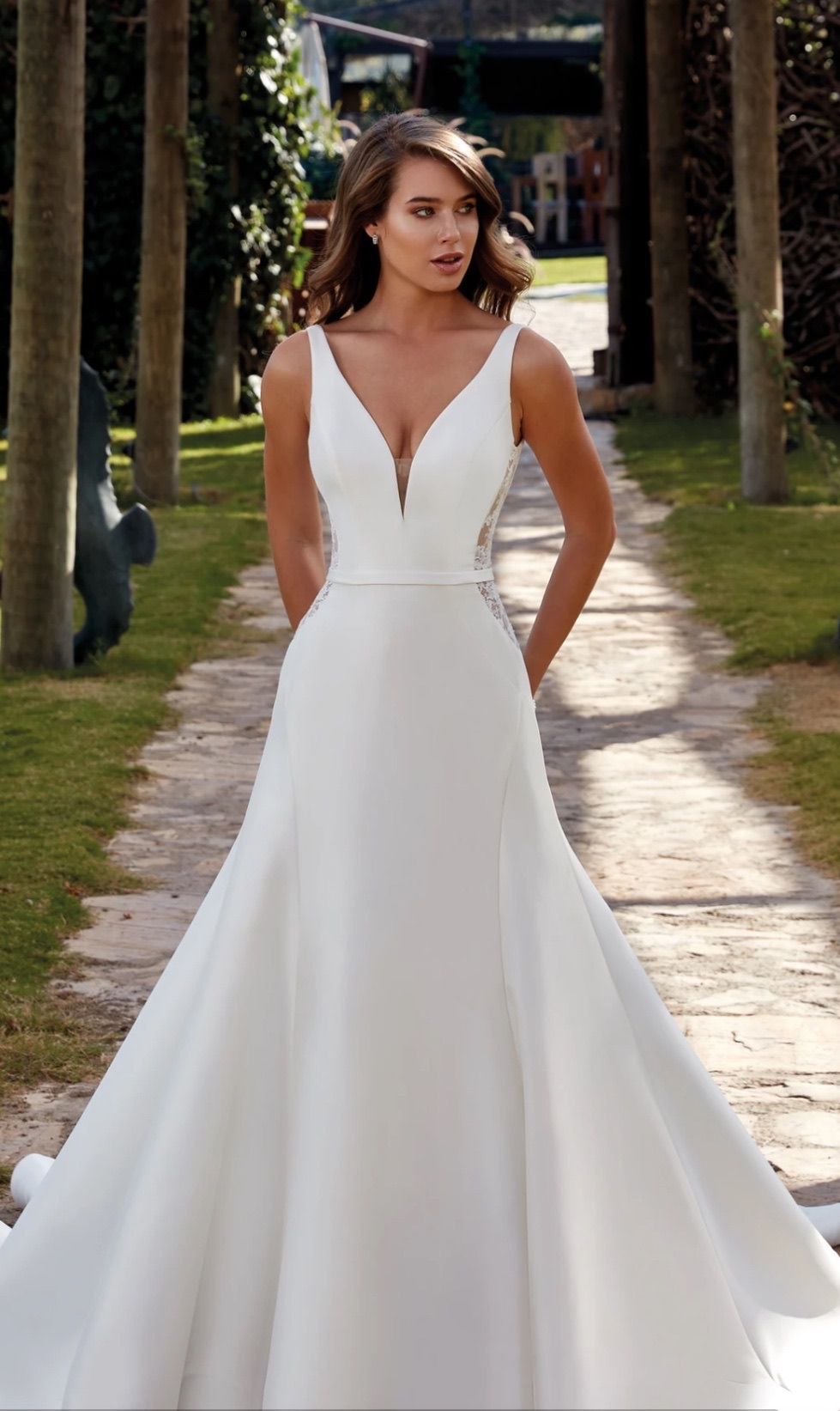 Woman in white wedding dress with deep V-neckline standing on a garden path with hands behind back.