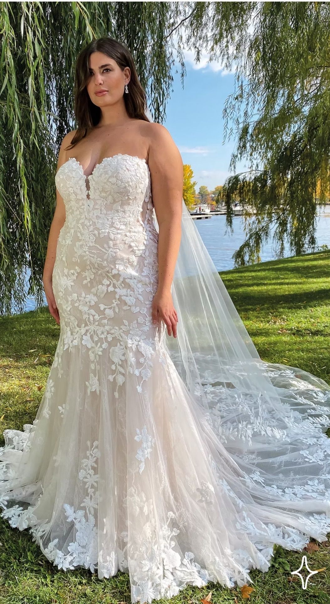 Woman in a strapless white lace wedding dress with a long train, outdoors near water.