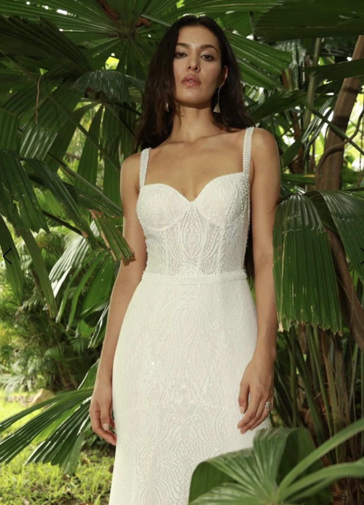Woman in a white beaded wedding dress with spaghetti straps, standing in front of tropical foliage.