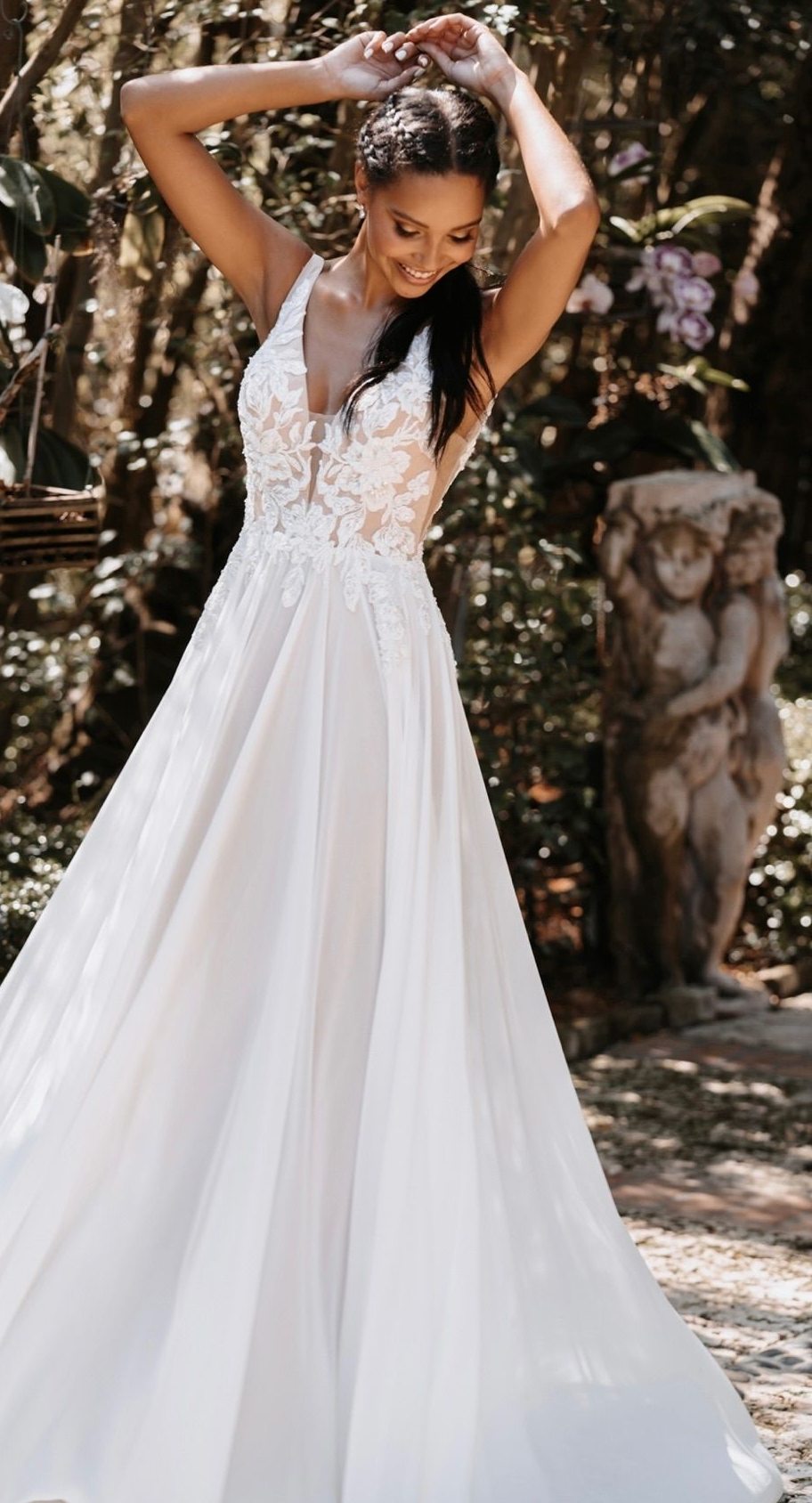 Woman in white wedding dress, arms raised, smiles. Outdoors, garden setting with statues.