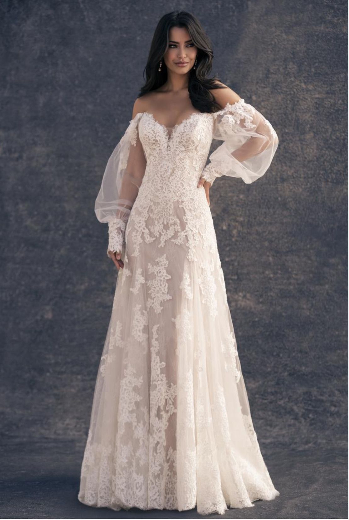 Woman in off-the-shoulder, lace wedding dress with sheer sleeves, posing against a dark background.