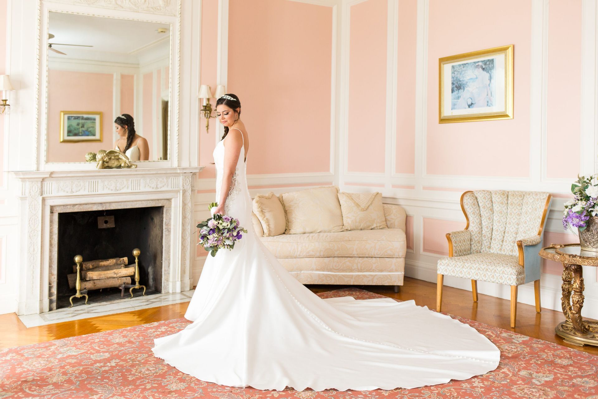 A bride in a long wedding dress is standing in front of a fireplace in a living room.