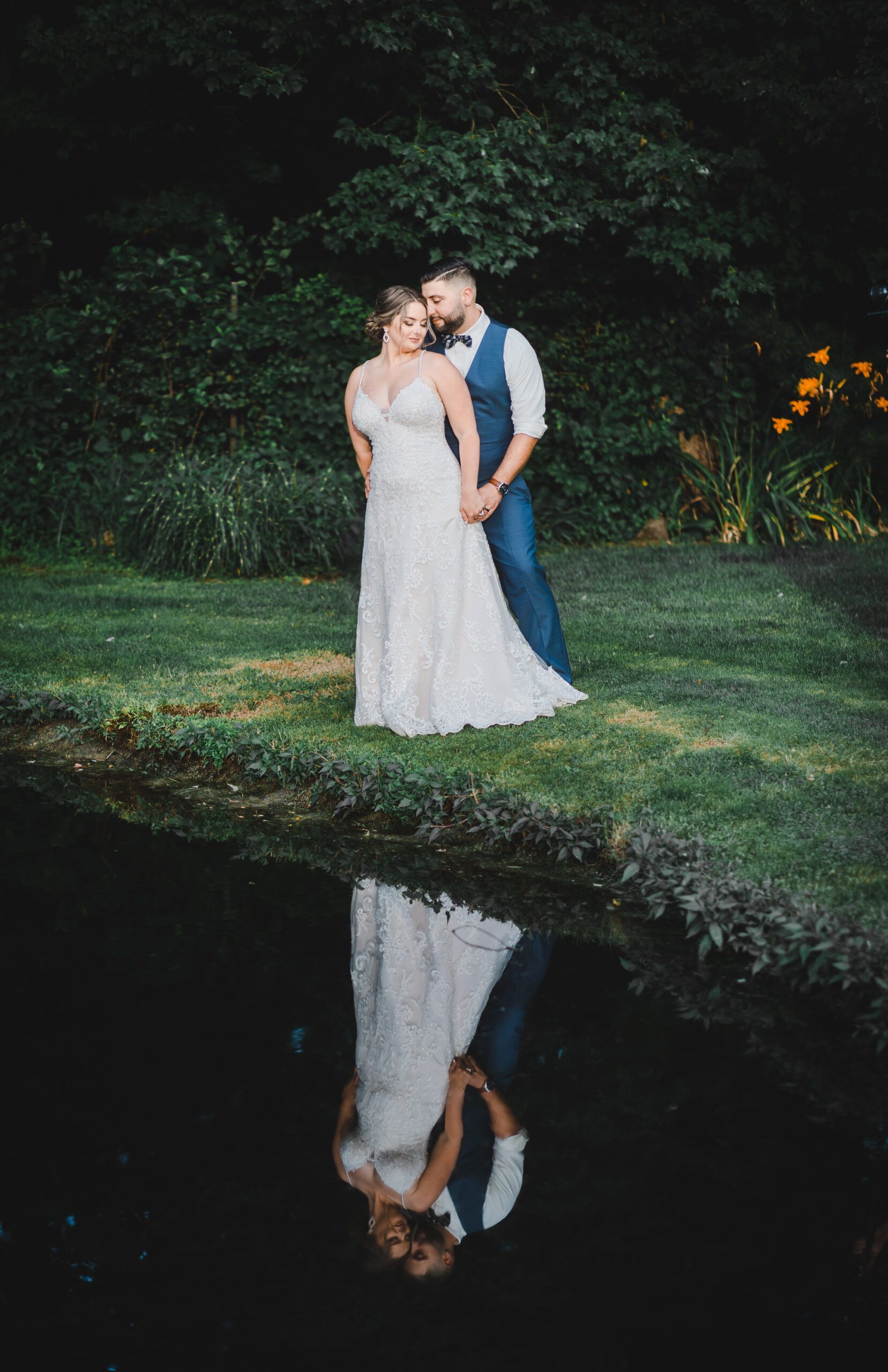 A bride and groom are standing next to a pond and their reflection is in the water.