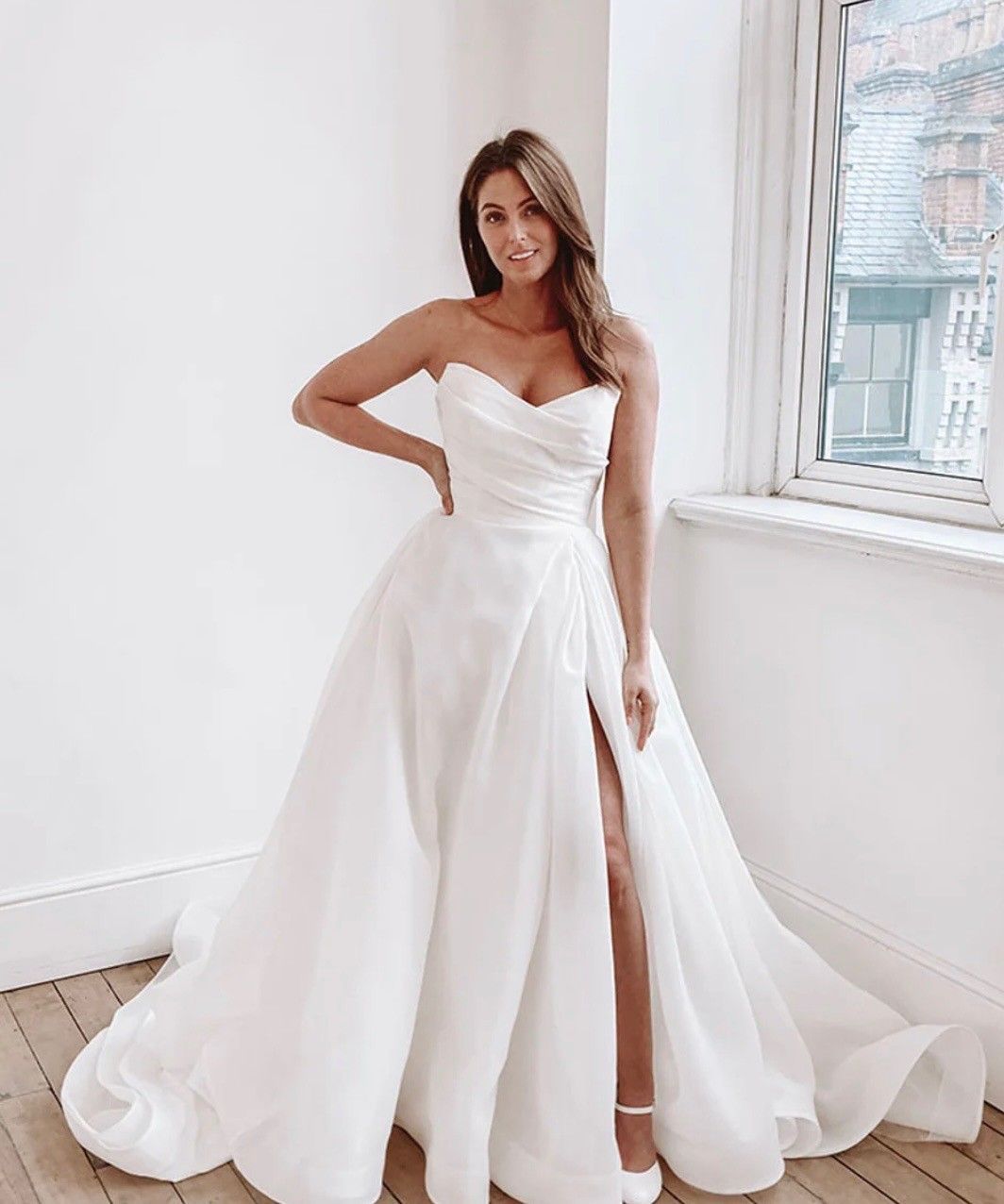 A person wears a strapless, white A-line gown with a thigh-high slit and pleated bodice, posing in a bright room.