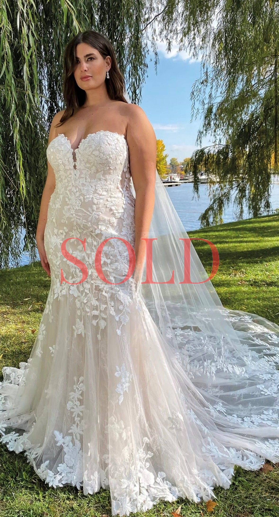 A person wearing a strapless, floral lace mermaid wedding dress with a long train, standing outdoors by a river.