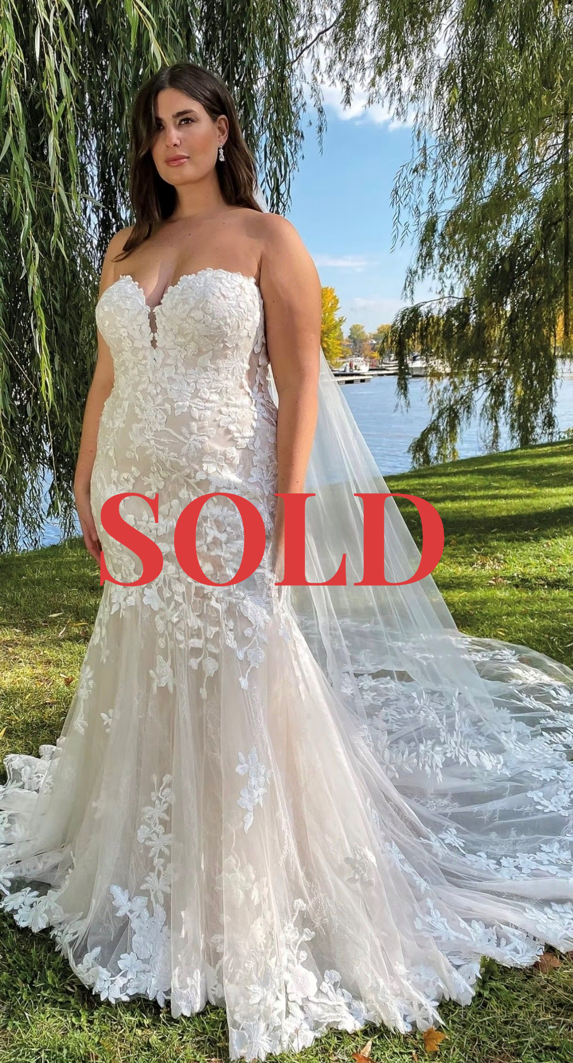 A person wearing a strapless, floral lace mermaid wedding dress with a long train, standing outdoors by a river.
