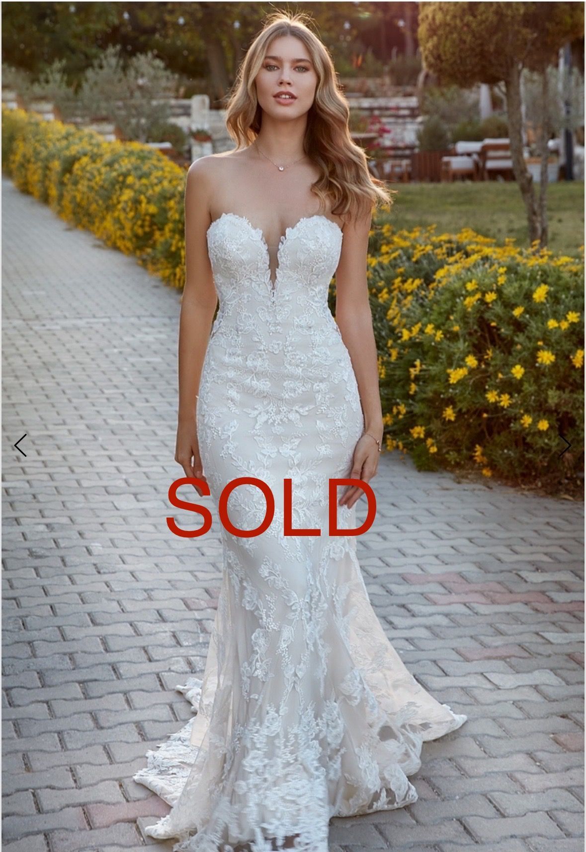 Woman in strapless white lace mermaid gown, posing on a brick pathway, surrounded by greenery and yellow flowers.