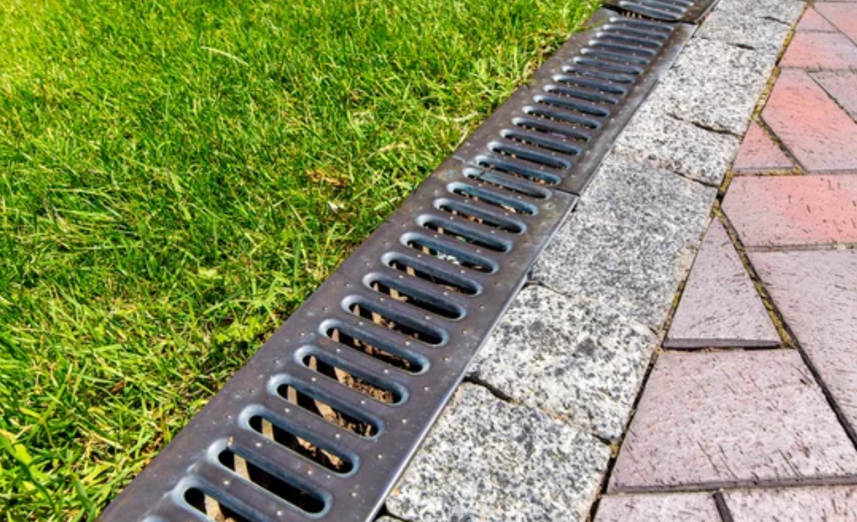 A metal drain is sitting on a brick sidewalk next to a lush green lawn.