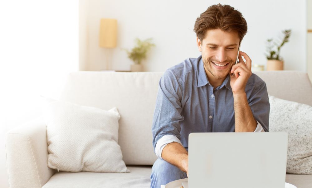 A man is sitting on a couch using a laptop and talking on a cell phone.