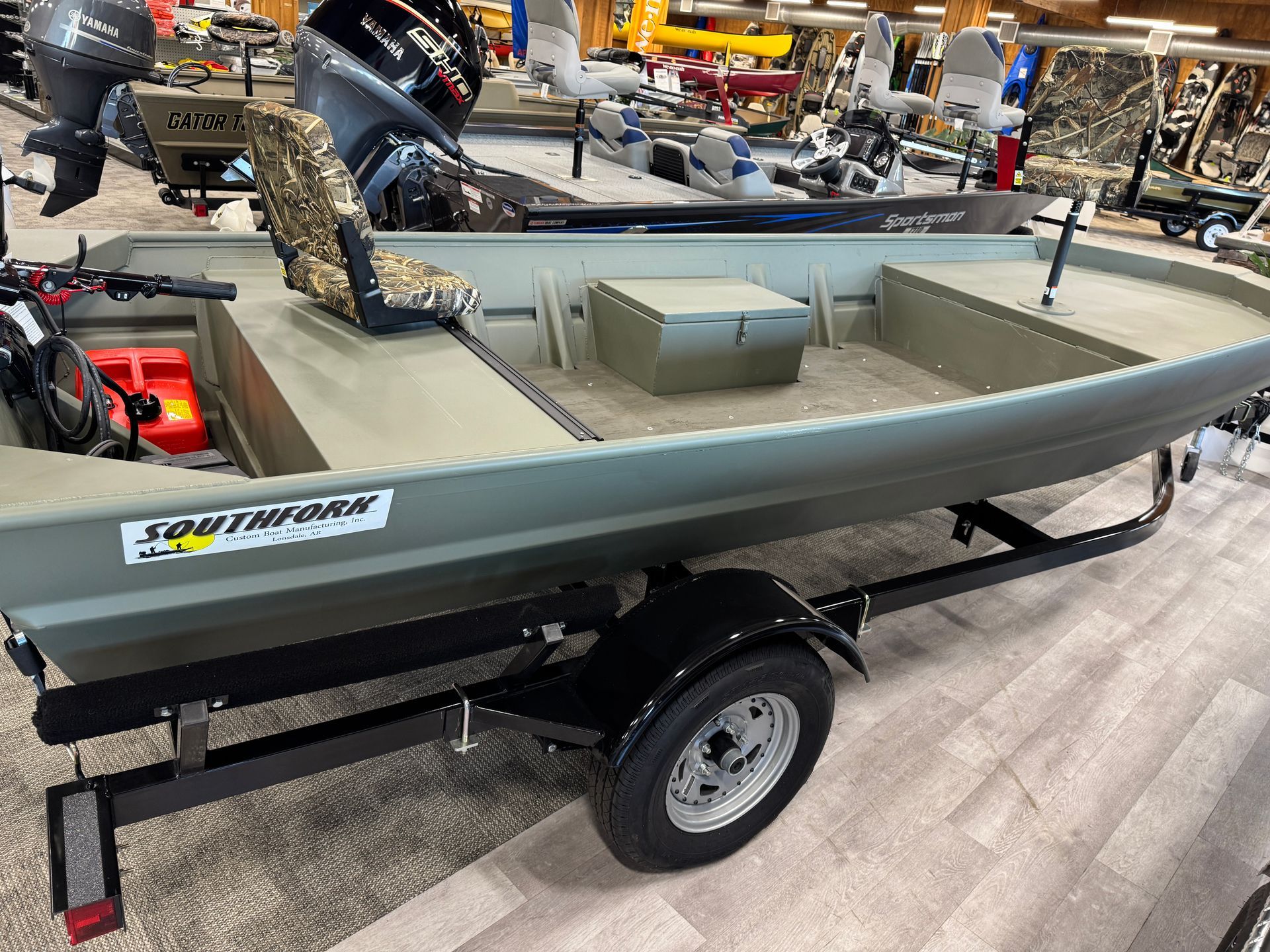 Olive green jon boat on a trailer, displayed indoors with other boats.