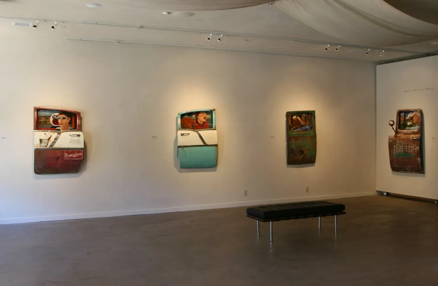 Art gallery with four wall-mounted, colorful pieces. A black bench sits in the foreground. White walls, bright lighting.