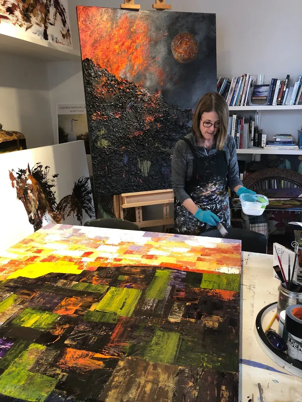 Artist in art studio, painting on large canvas with brush, surrounded by artwork and supplies.