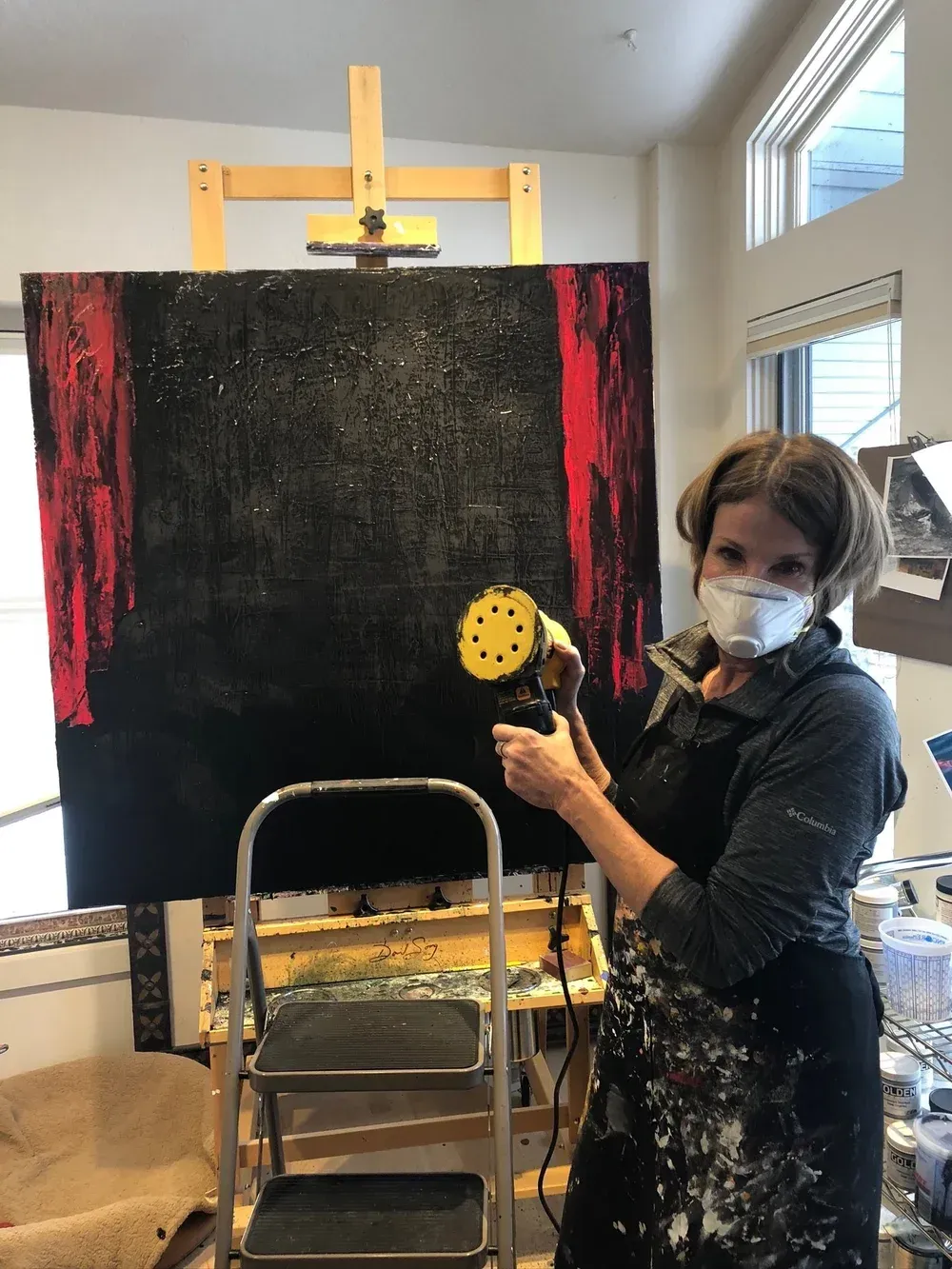 Woman sanding a large black and red abstract painting on an easel, wearing a respirator, in a studio.