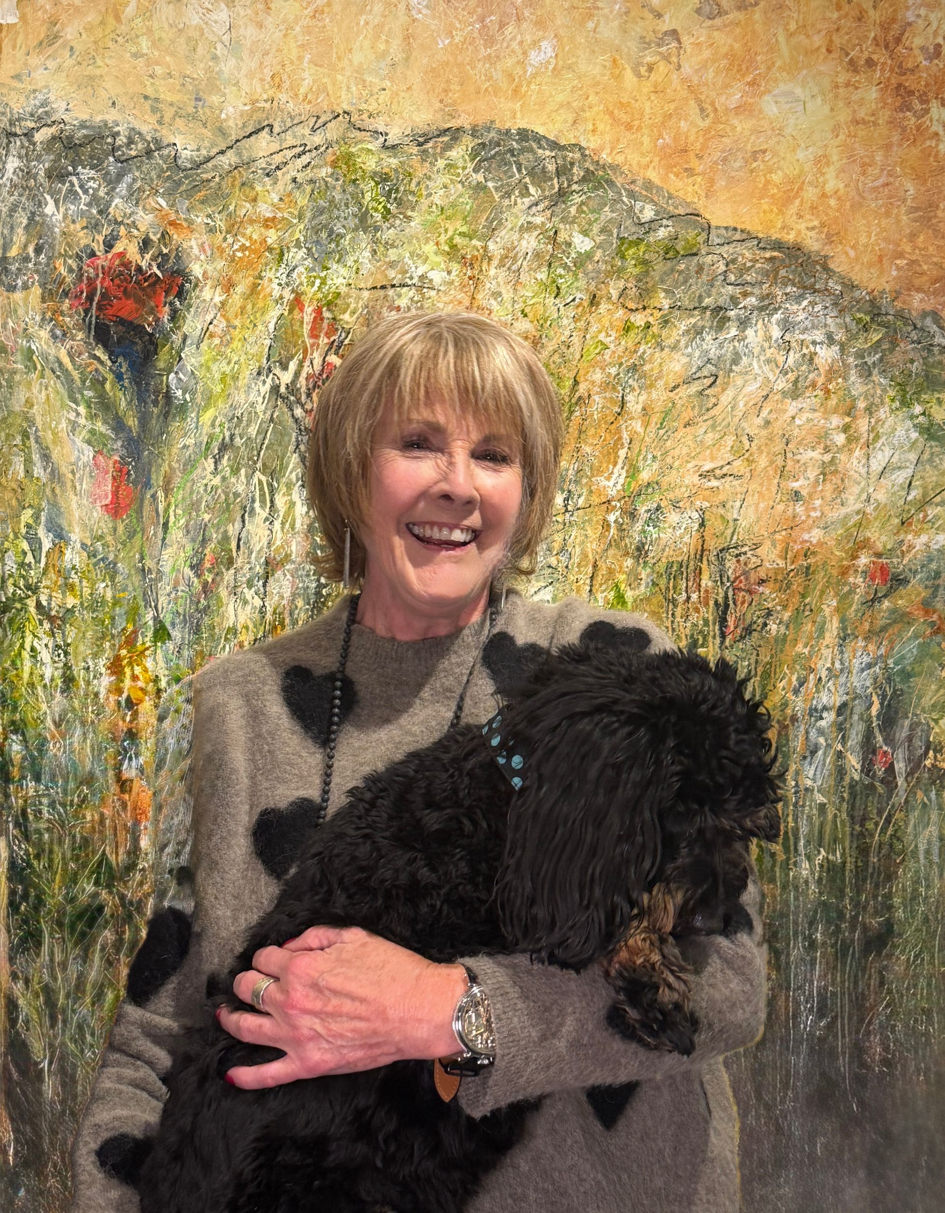 Woman smiling, holding a small black dog, stands in front of a colorful, impressionistic painting.