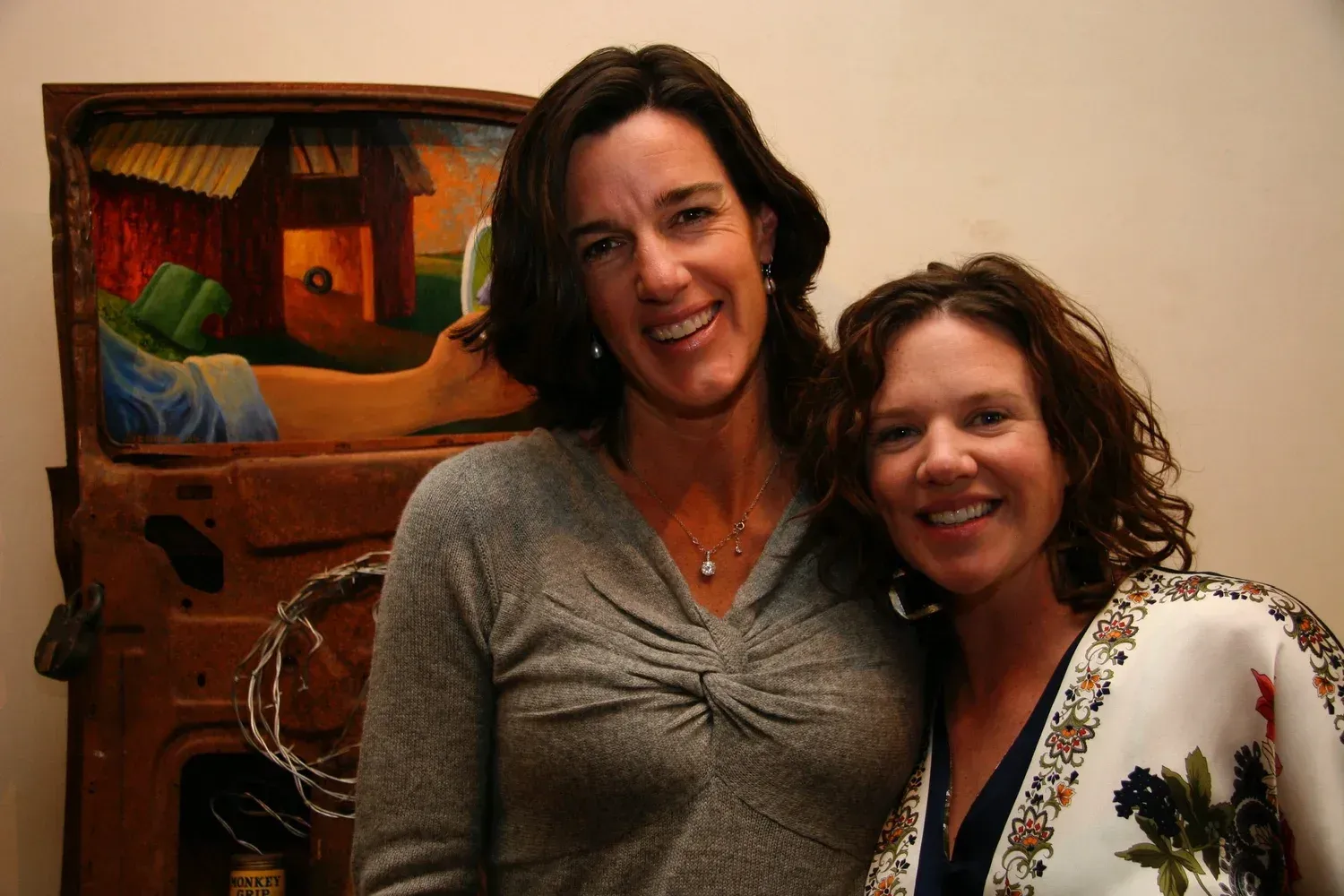 Two smiling women pose near art with rustic barn imagery.