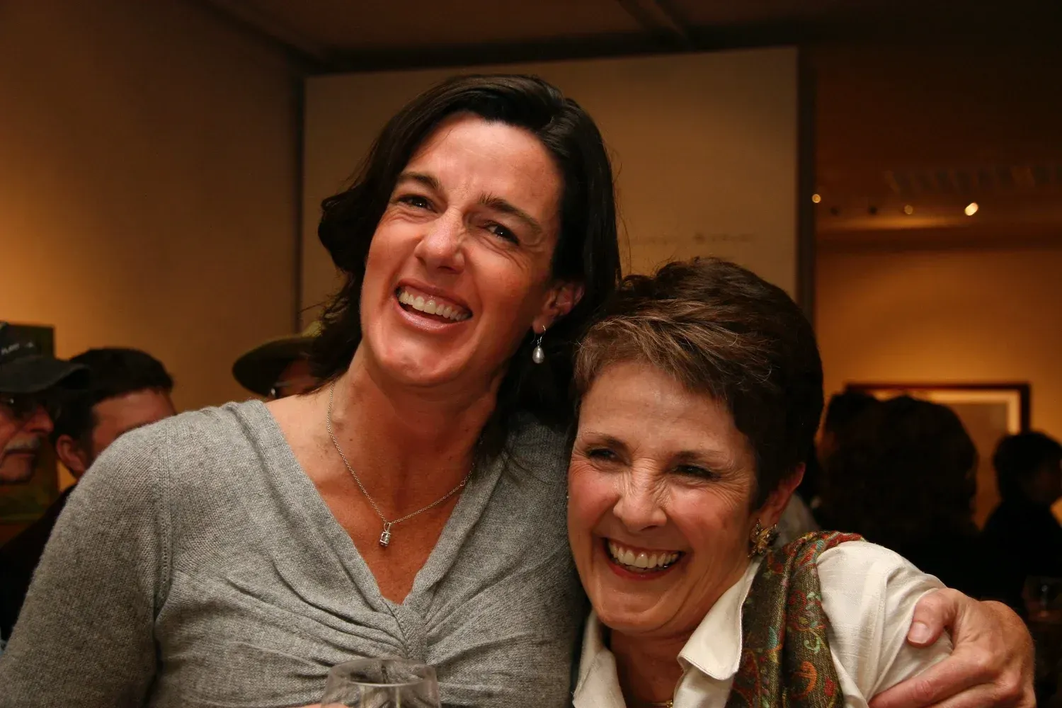 Two smiling women, one with short hair, arm around the other wearing a gray sweater, at an event.